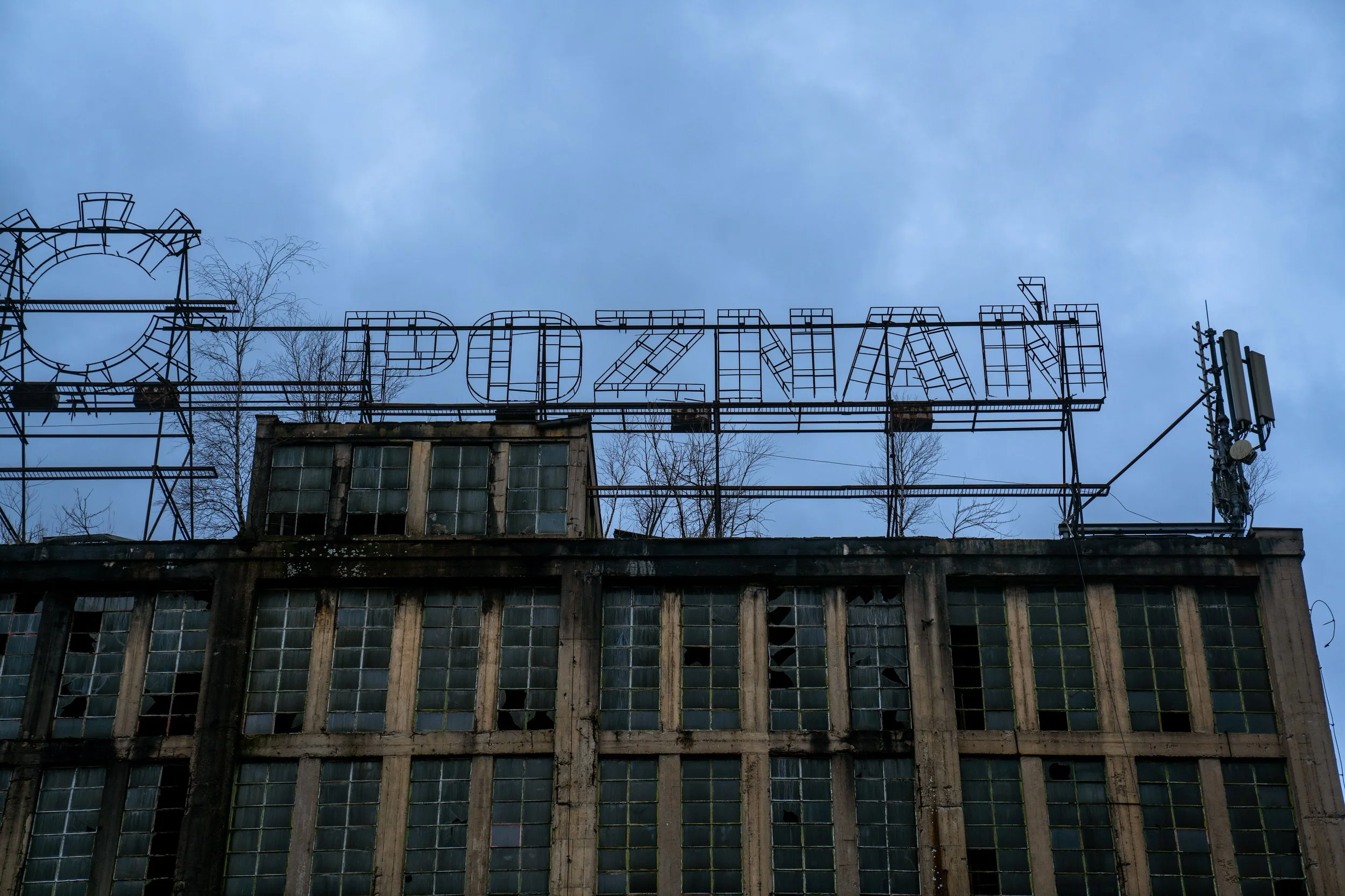 Old, abandoned building with broken windows and a rusted sign reading 'POZNAŃ' on the roof, set against a cloudy sky.