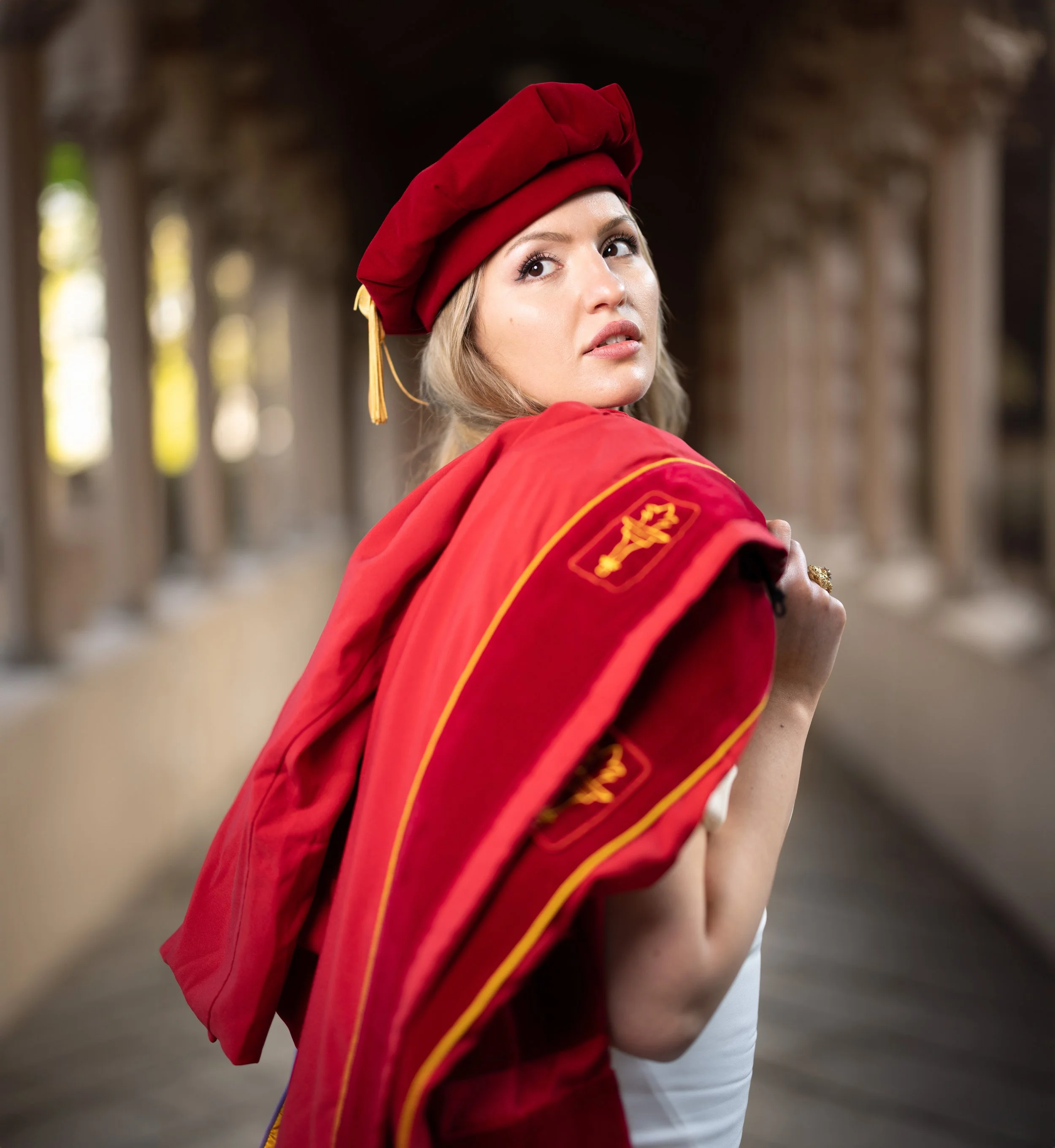 A young woman dressed in a traditional graduation cap and gown holding the gown over her shoulder, standing in a historic corridor with arches and columns.