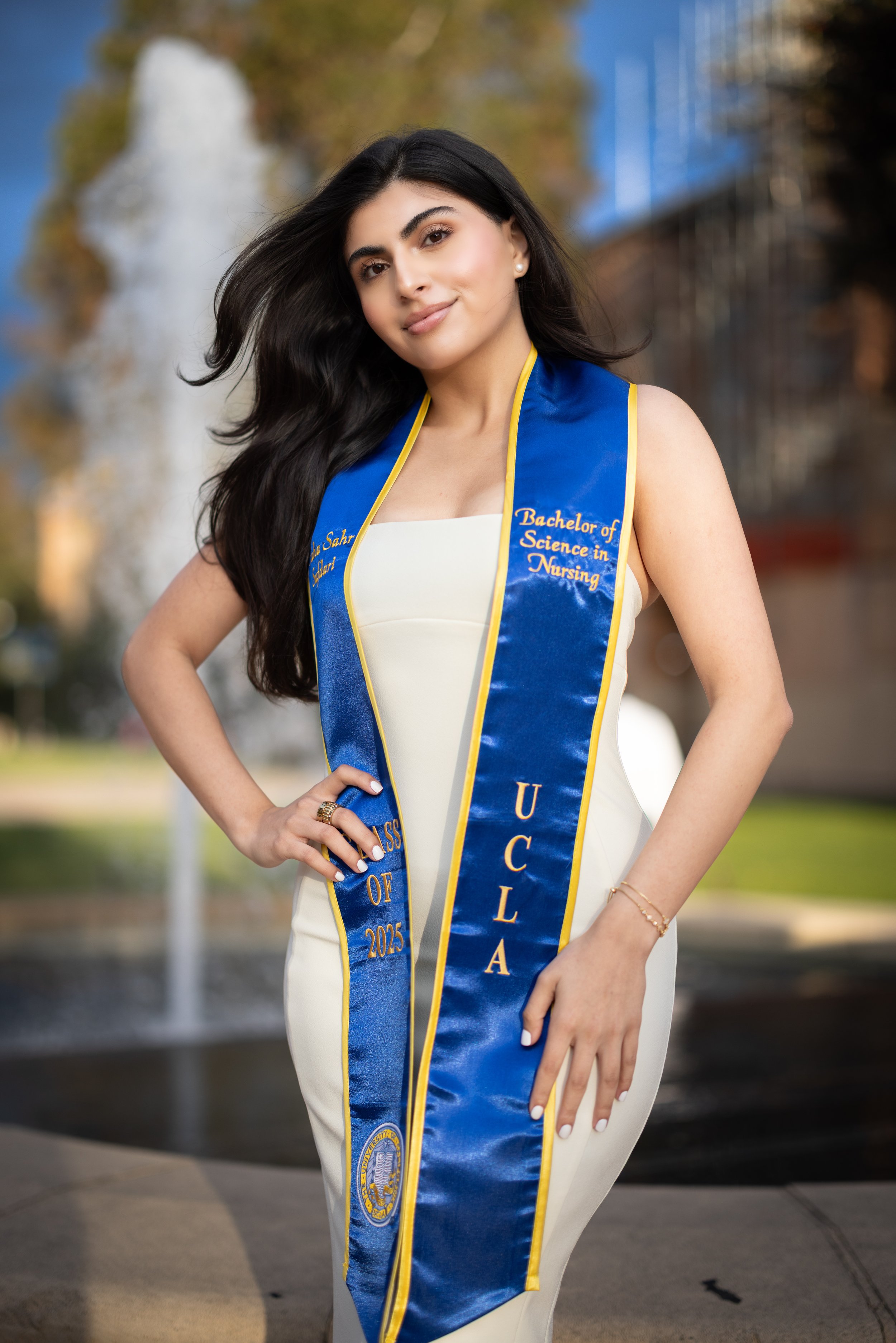 A young woman with long dark hair and light skin wearing a white dress and a blue graduation sash from UCLA, celebrating her bachelor of science in nursing degree in 2023, standing outdoors near a fountain with trees and buildings in the background.
