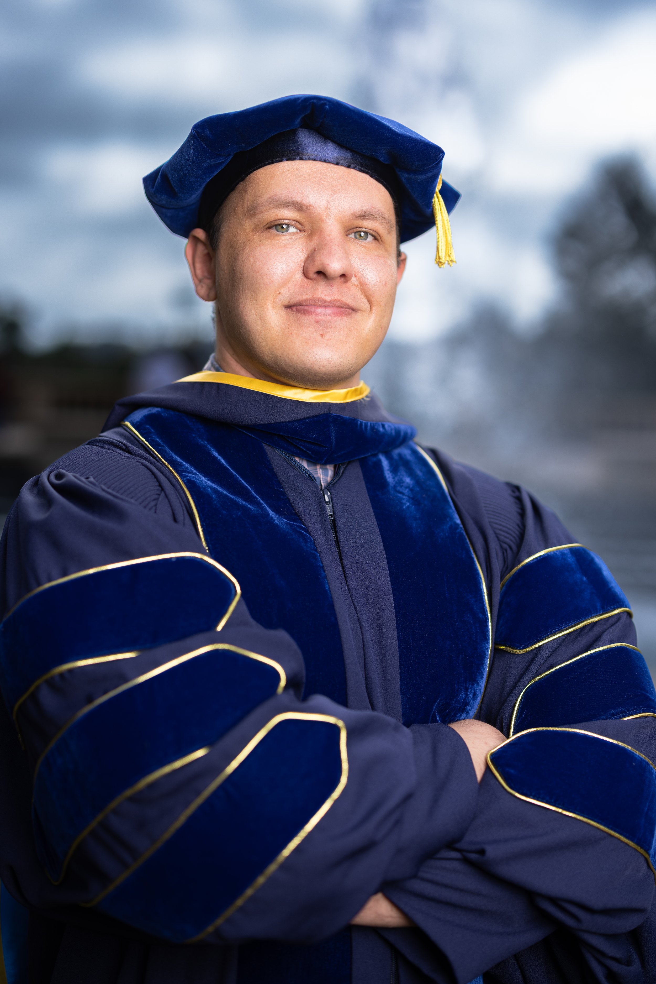 Young man wearing academic regalia, including a blue and gold gown and a blue cap with a yellow tassel, standing outdoors with arms crossed and smiling.