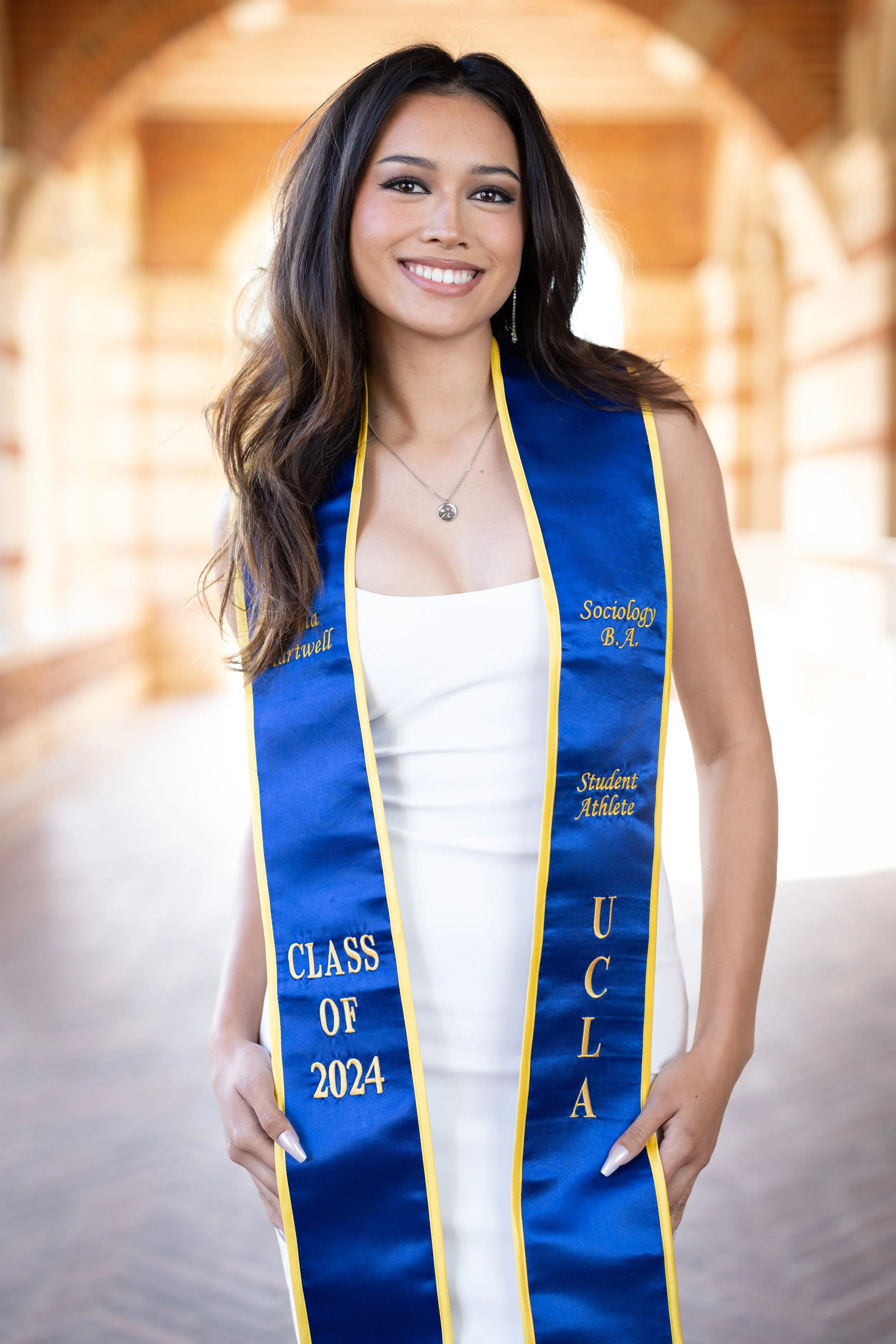 A young woman wearing a white dress and a blue and yellow graduation sash that reads 'Class of 2024,' 'UCLA,' 'Sociology B.A.,' and 'Student Athlete,' standing in a well-lit indoor space with arched windows.