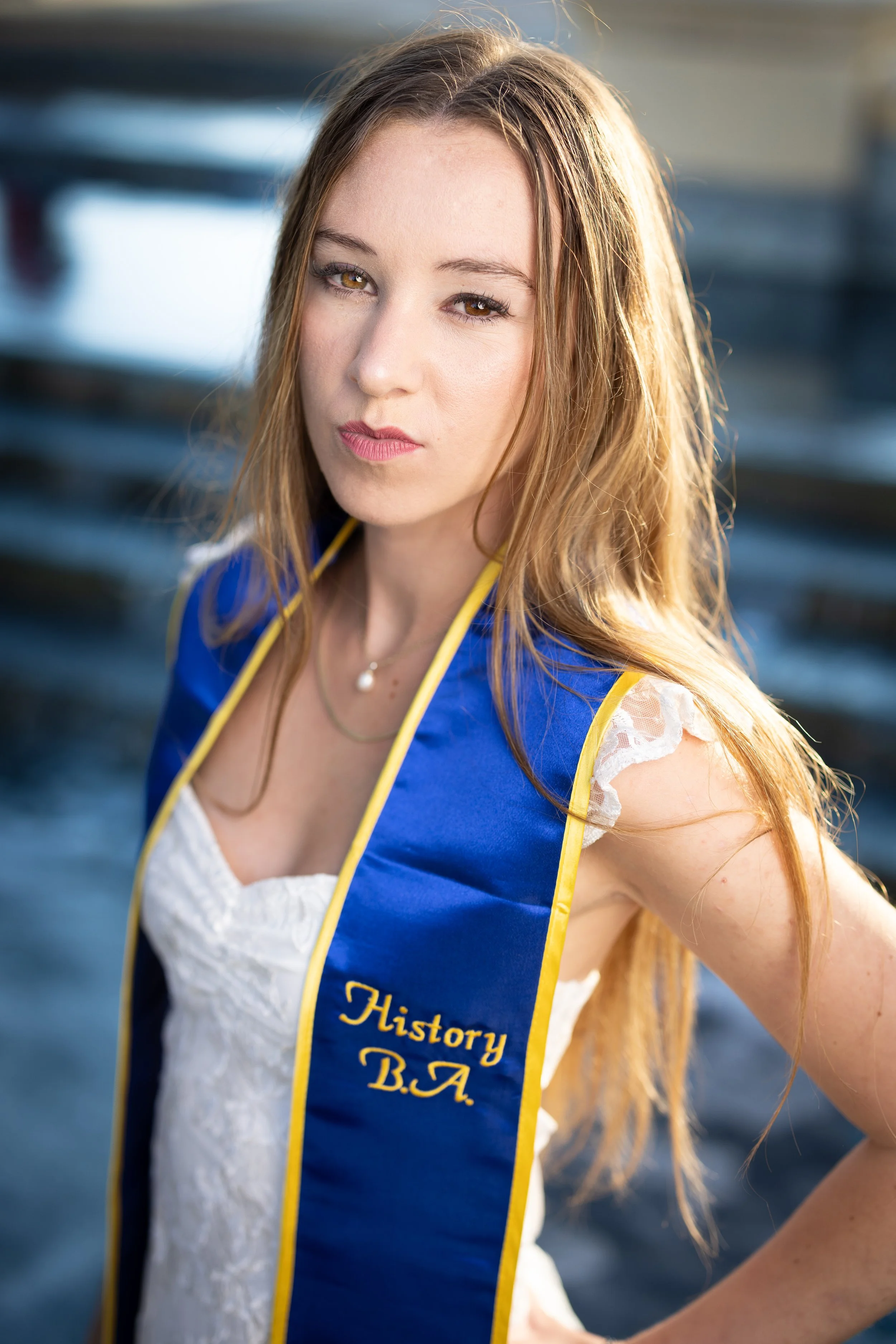 A young woman with long, light brown hair and a serious expression, wearing a blue and yellow sash that reads 'History B.A.' over a white dress, outdoors with blurred background of water and boats.