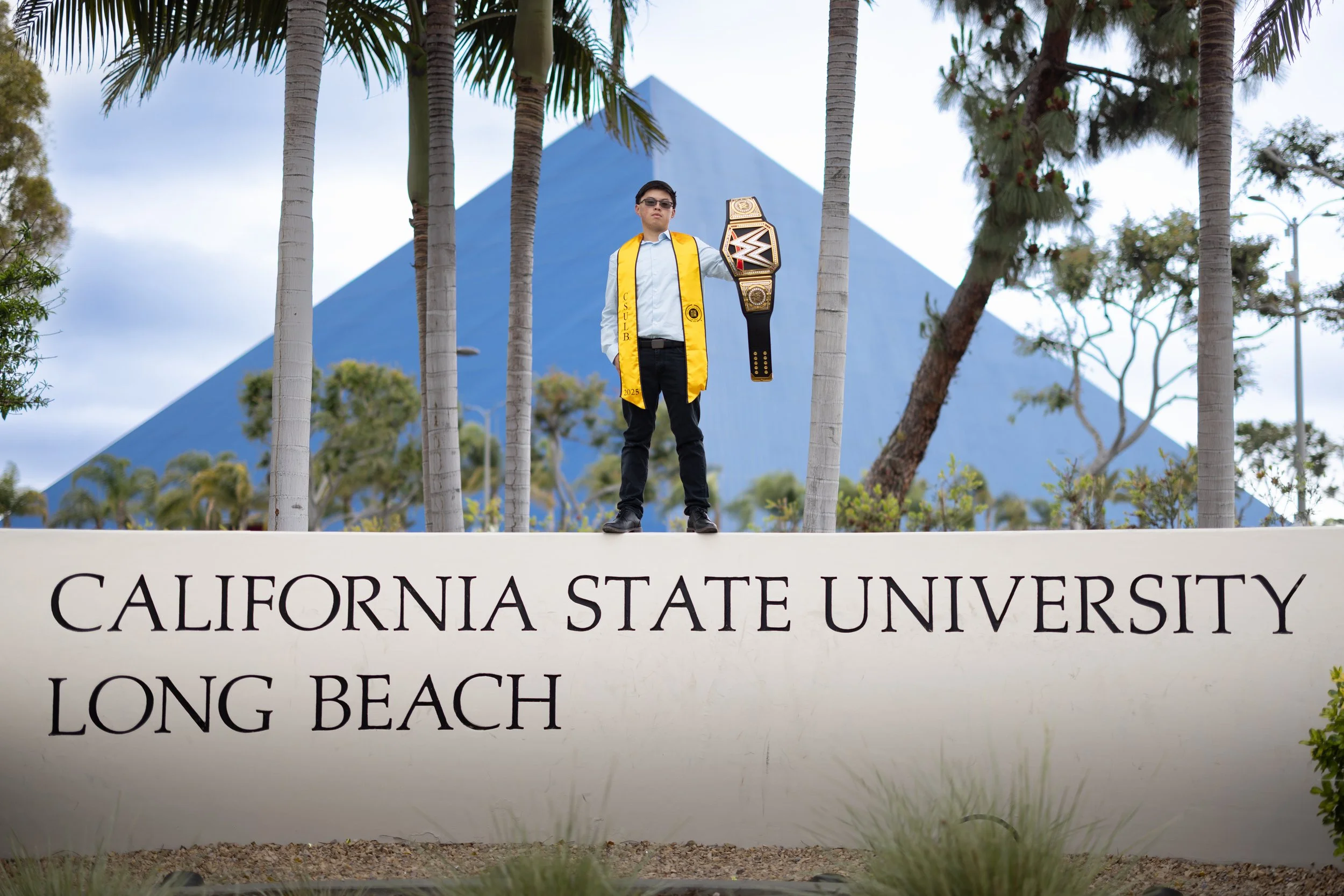 A young man in a light blue shirt, black pants, and sunglasses, wearing a yellow graduation stole, stands on a sign that reads 'California State University Long Beach.' He is holding a WWES championship belt, with a large, pyramid-shaped building and palm trees in the background.