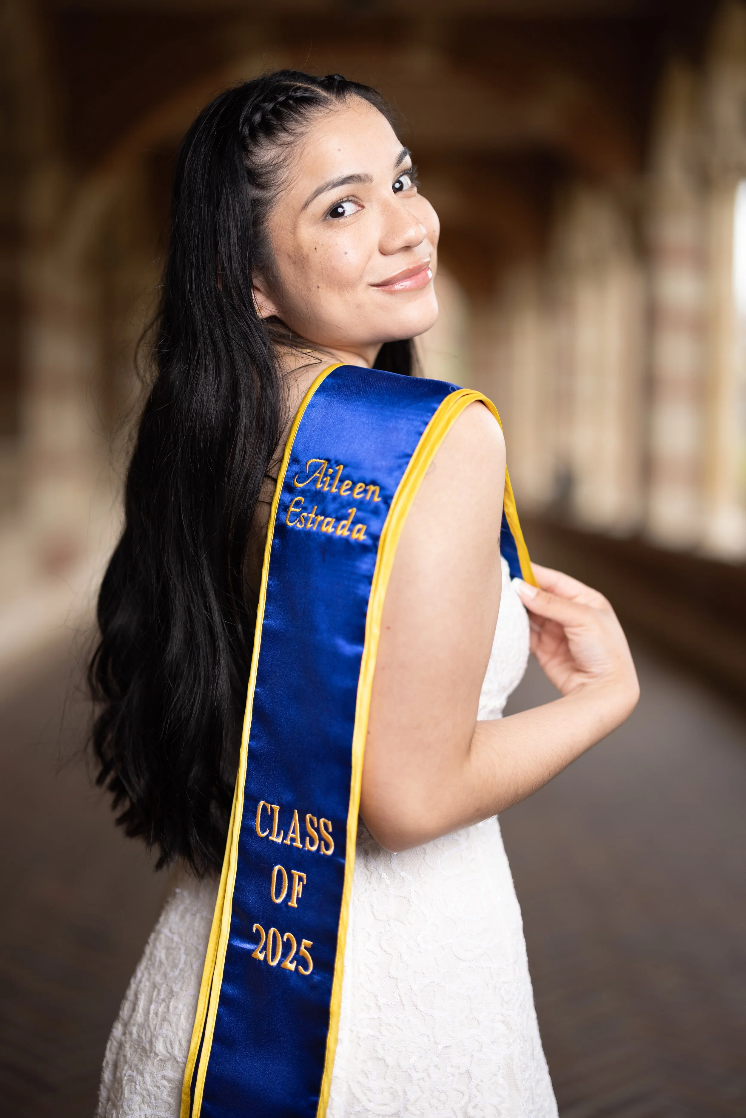 A young woman with long dark hair, wearing a white dress and a blue sash with yellow trim that reads "Aileen Estrada, Class of 2025," standing inside a covered outdoor walkway and smiling at the camera.