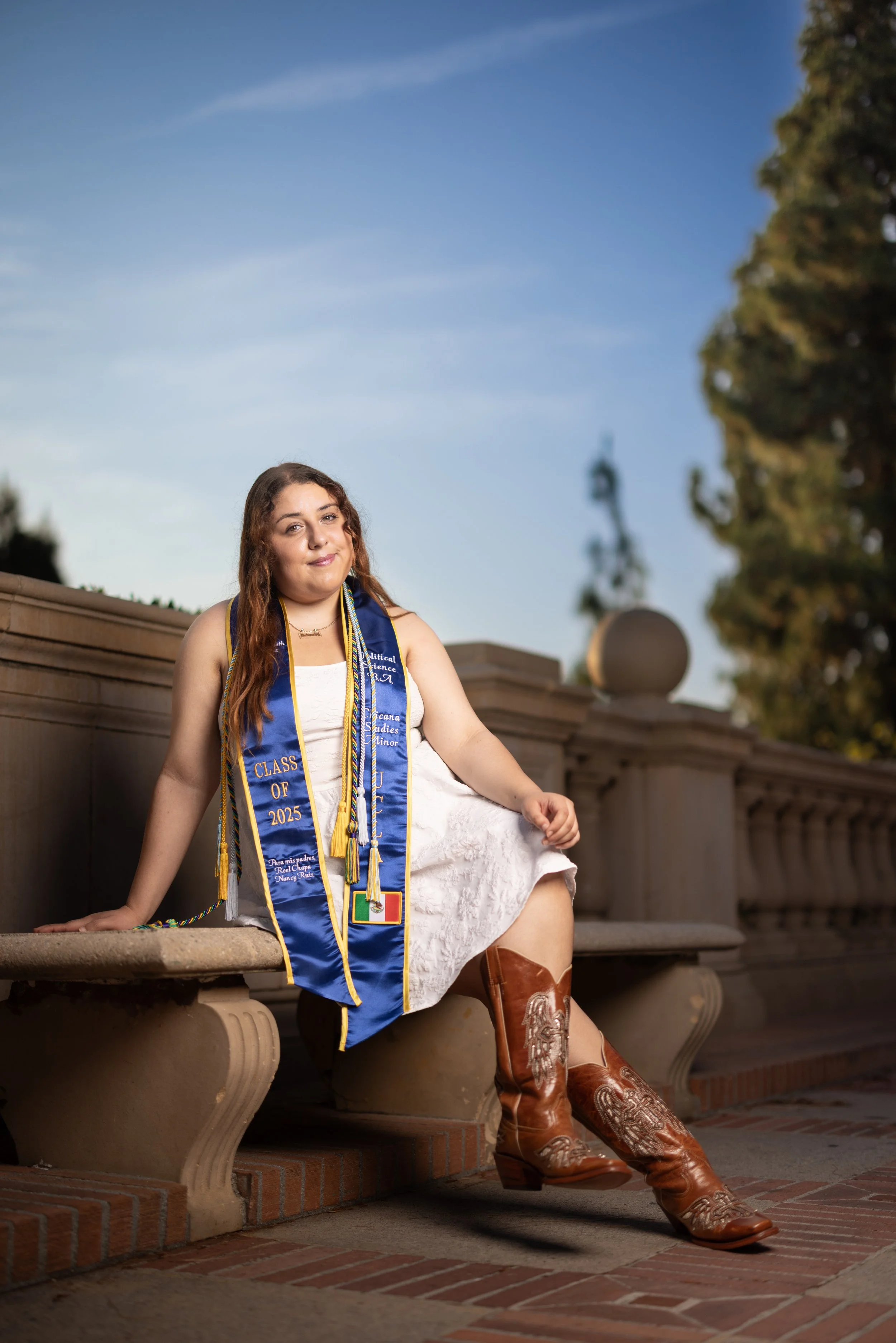 A young woman sitting on a stone bench outdoors during the daytime. She is wearing a white dress, cowboy boots, and a blue graduation stole with gold accents that reads "Class of 2025." She has long, wavy brown hair and is smiling softly. The backgro