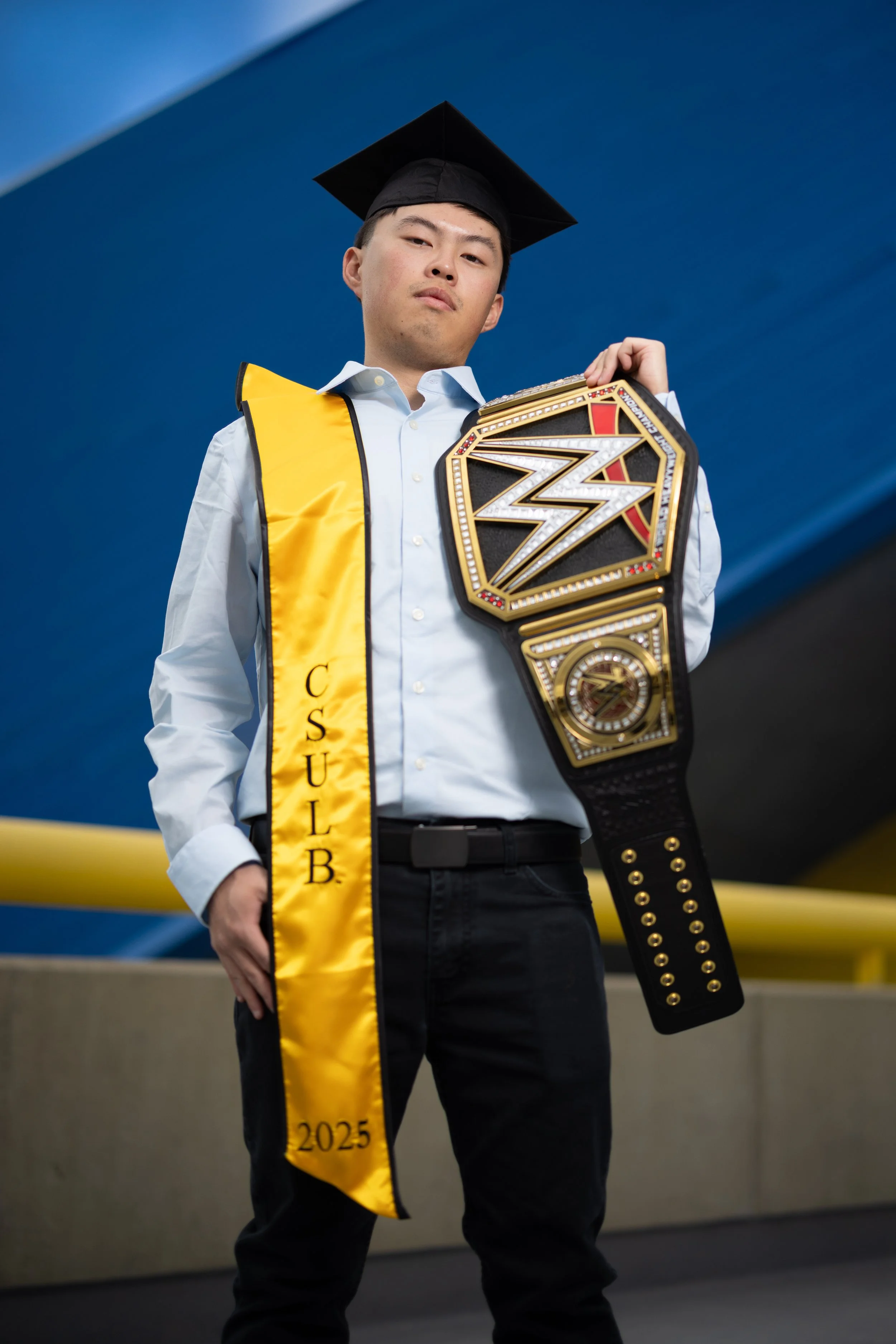 Young man in a white shirt and black pants wearing a graduation cap and sash, holding a WWE championship belt.