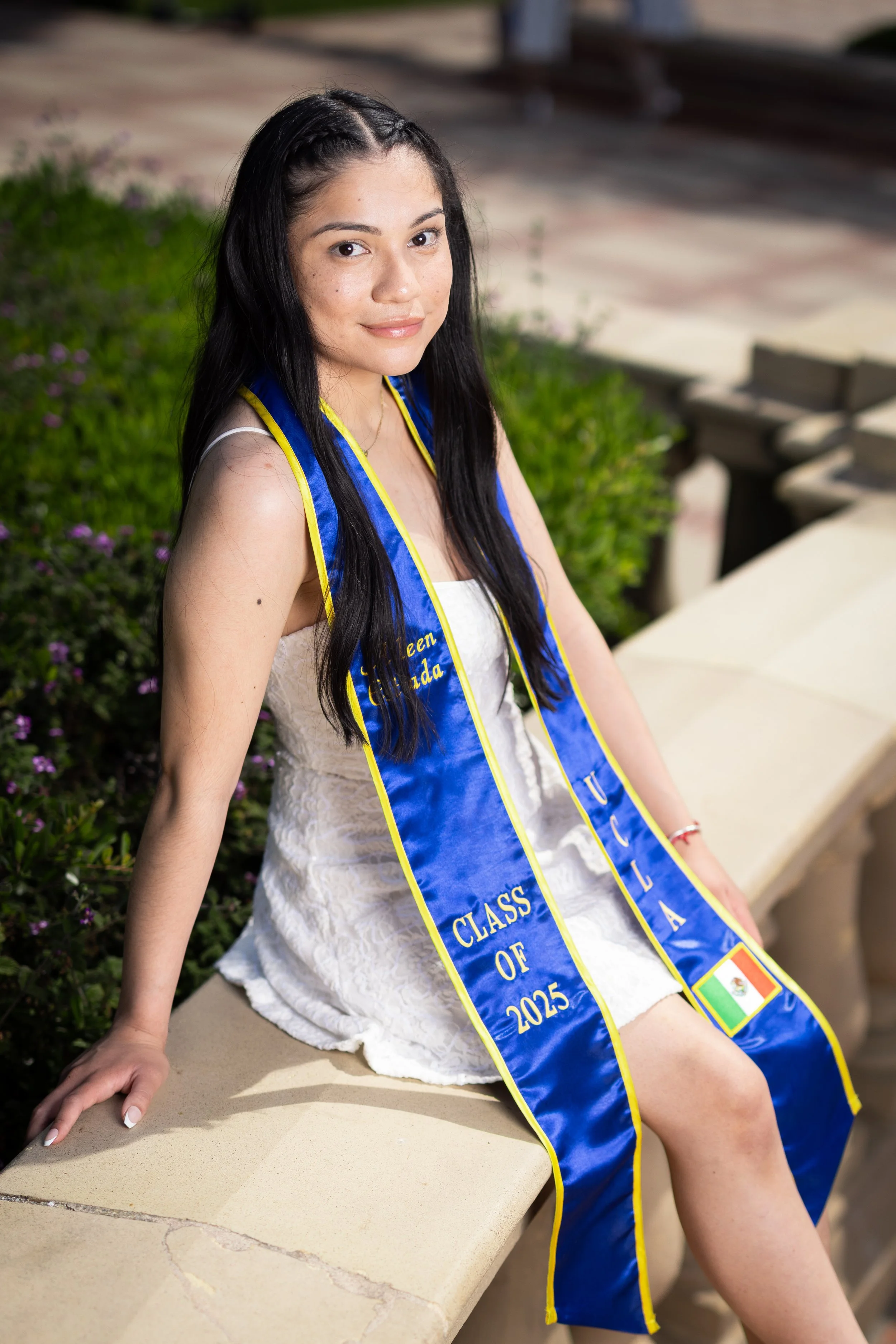 Young woman wearing a blue graduation stole with yellow trim, sitting outdoors on a stone bench, surrounded by greenery and purple flowers.