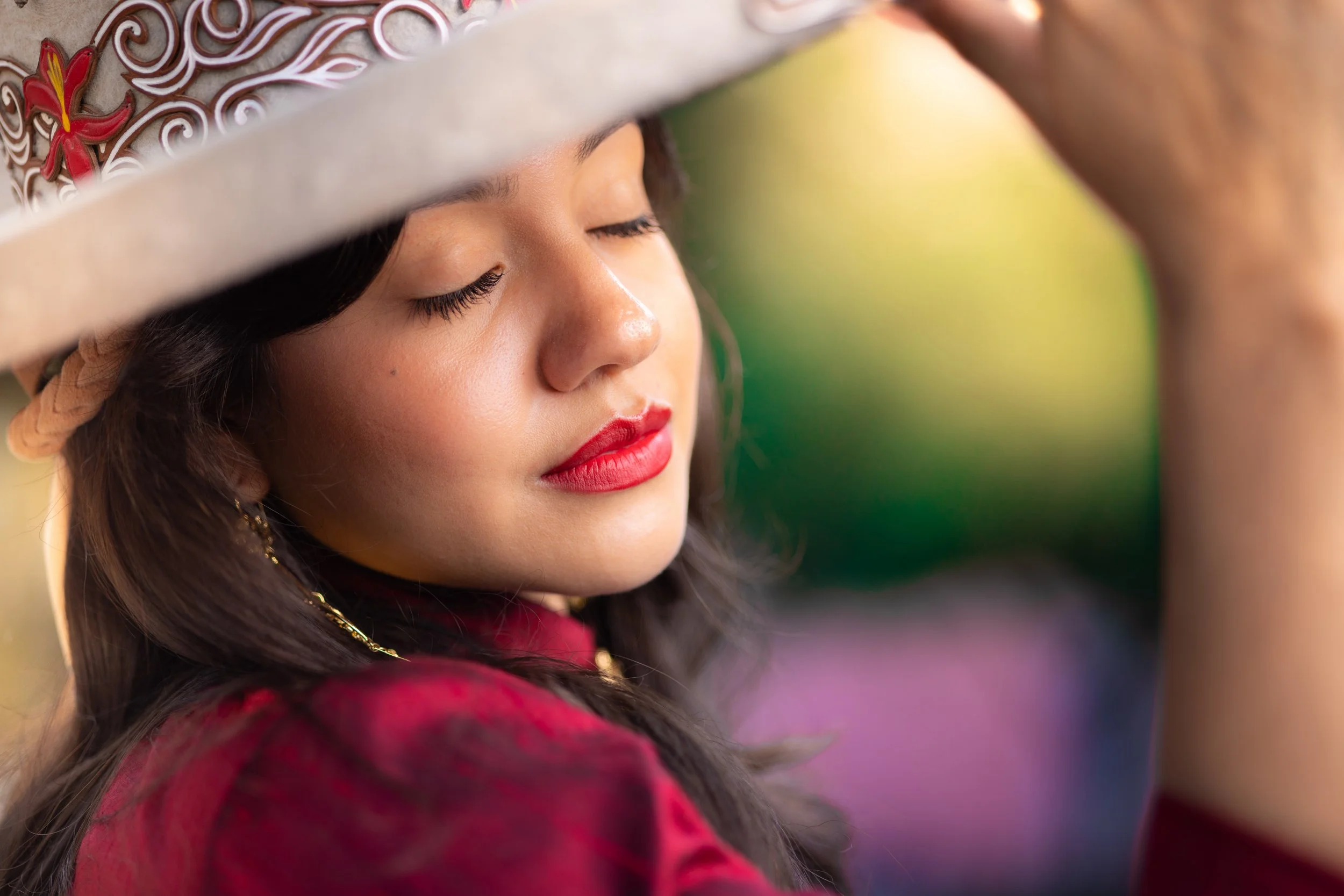 A woman wearing a traditional hat with embroidery and a red outfit, with her eyes closed and leaning her head slightly, showing her makeup and earrings.