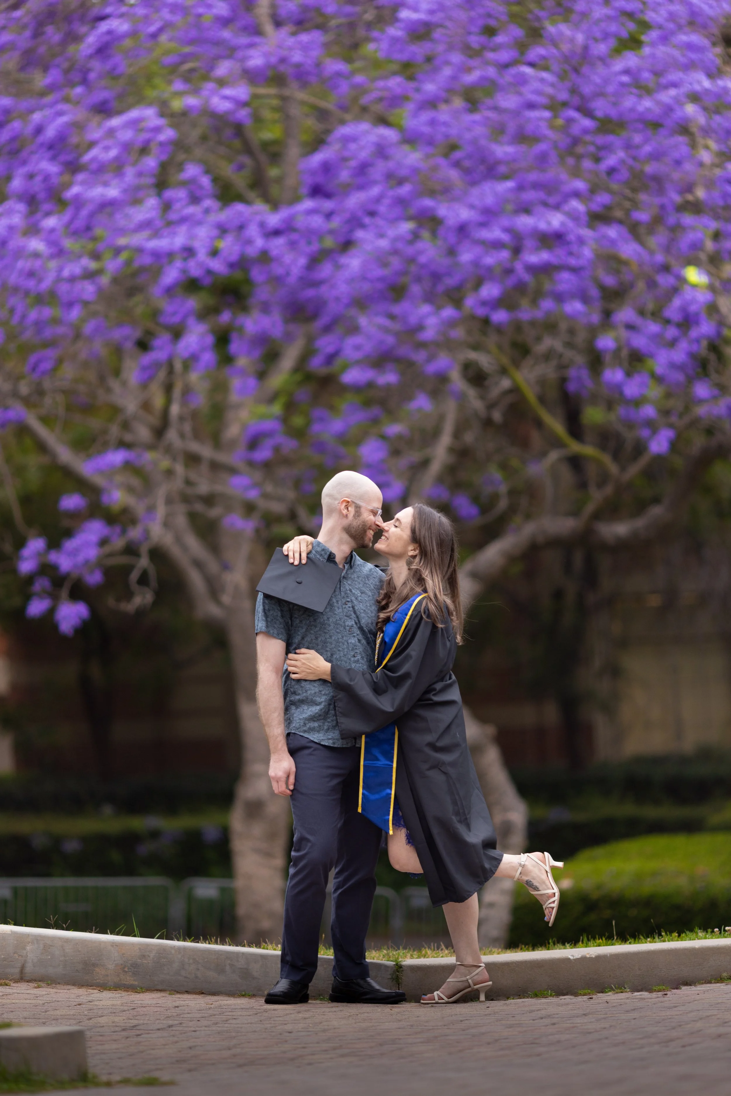 A couple celebrates graduation outdoors with purple flowering trees in the background, embracing and smiling at each other.