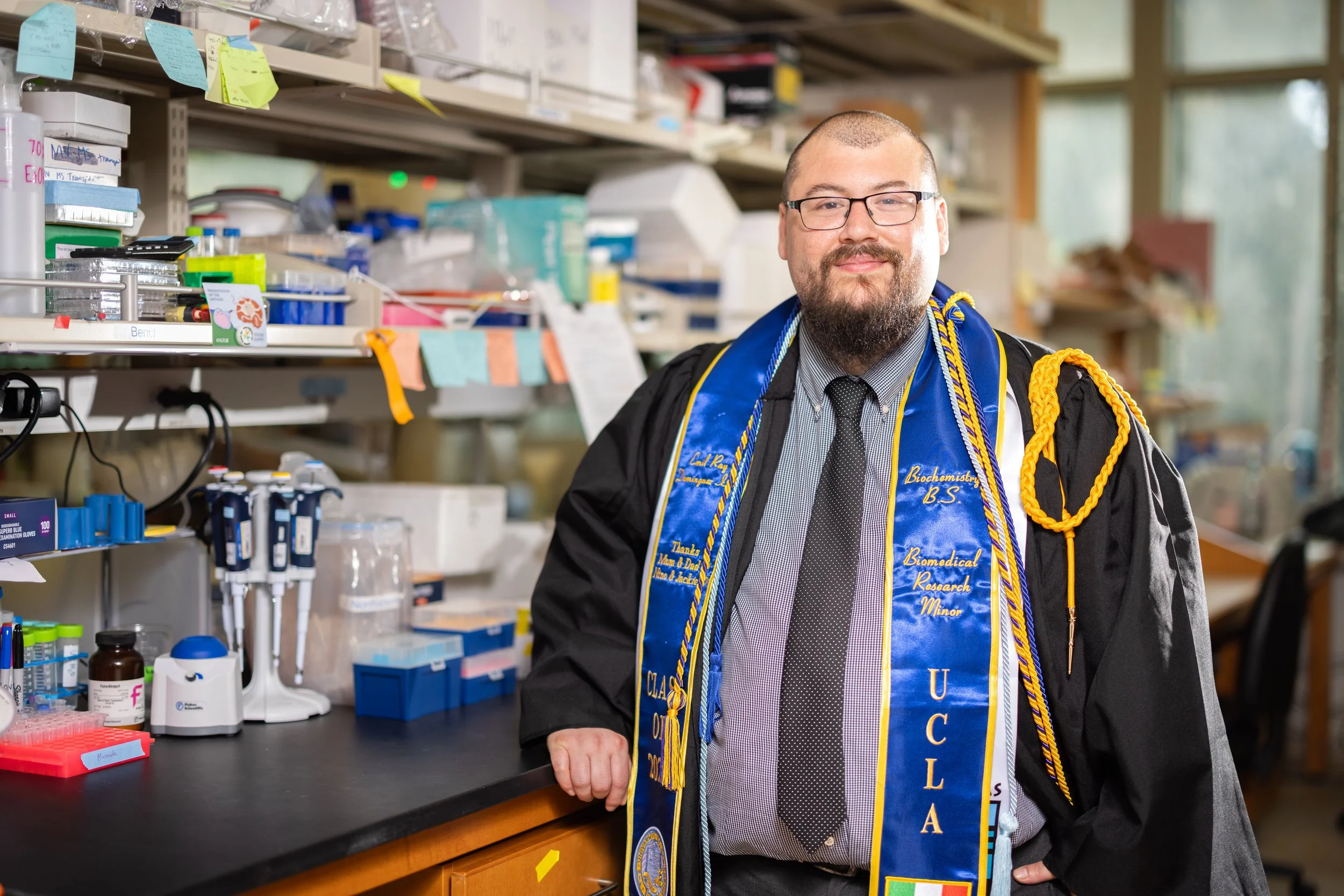 A man dressed in a graduation gown and honor cords standing in a laboratory surrounded by scientific equipment.