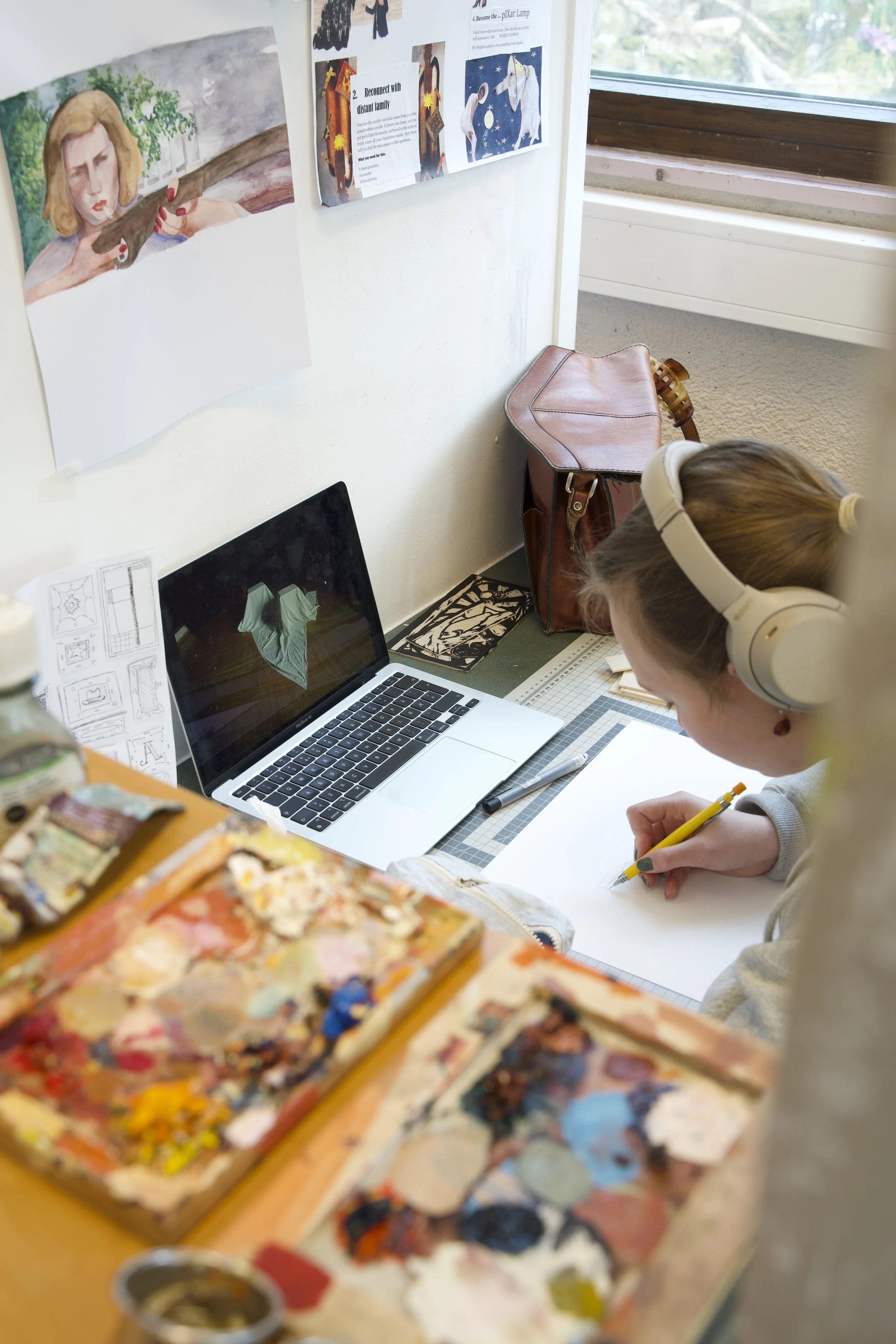 En jente med blond hår og hvite hodetelefoner som skriver med en gul penn på et hvitt ark, på et arbeidsbord med en kunstnermaling i forgrunnen og en laptop, veggpynt og et vindu bak.