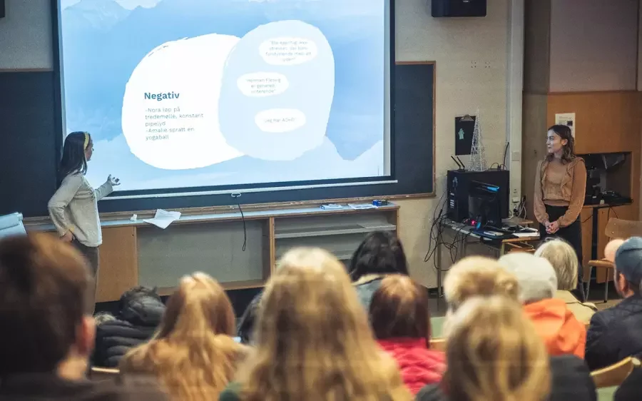 En gruppe mennesker i et klasserom ser på to kvinner som holder en presentasjon foran en stor skjerm med en PowerPoint-lysbilde. Publikum ser ut til å være elever eller studenter.