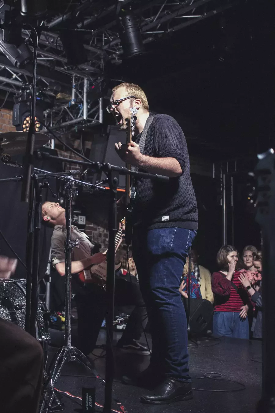 En musiker som spiller gitar på en konsert, omgitt av annet musikkutstyr og publikum i bakgrunnen.