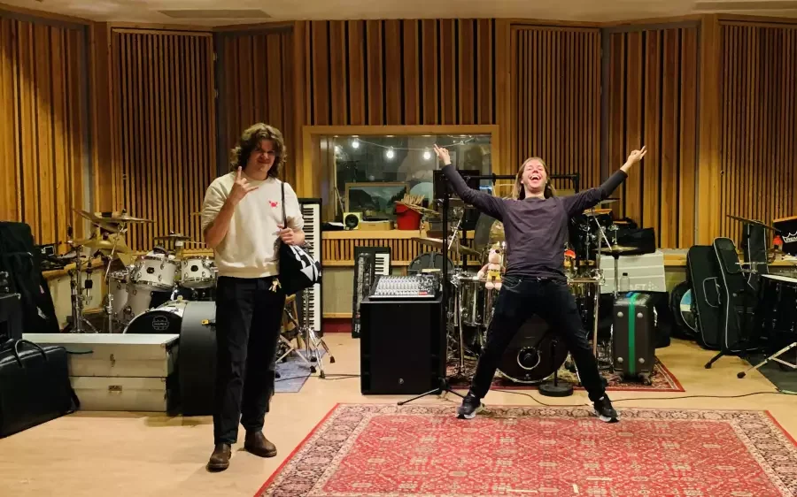 To personer i et lyd studio, den ene er ung mann med langt hår i en slags hvit genser, den andre er ung kvinne med kort hår og svart klær, hun smiler og har armene ut til siden.