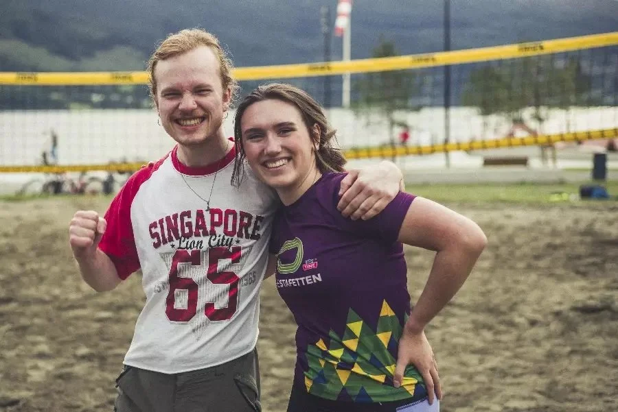To personer smiler og holder rundt hverandre på en sandvolleyballbane, med volleyballnett i bakgrunnen.