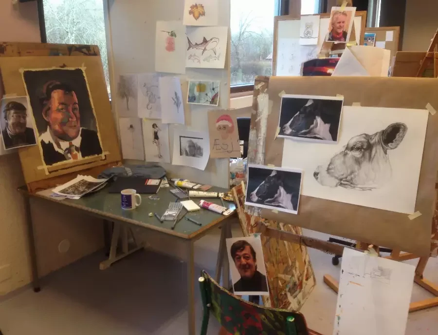 Et kunstverksted med flere portrett- og dyretegninger og malerier på veggen, samt maling og kunstutstyr på bordet.