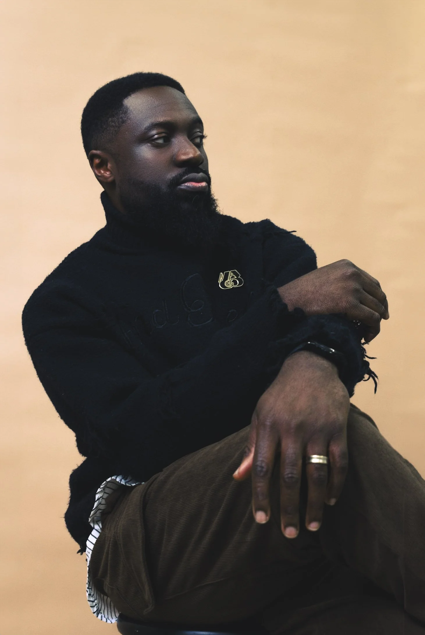 Person with a beard in a black turtleneck sweater, wearing a gold ring, sitting against a beige background.