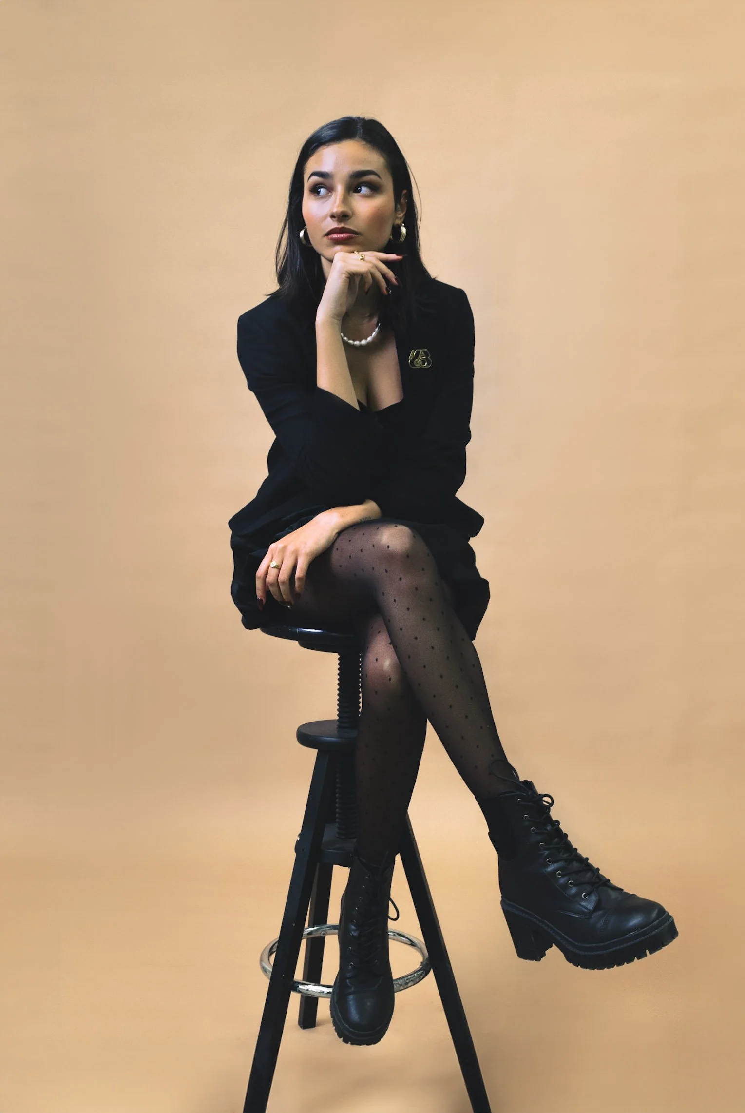 Woman sitting on a stool wearing a black outfit, polka dot tights, and black platform boots, posed against a beige background.