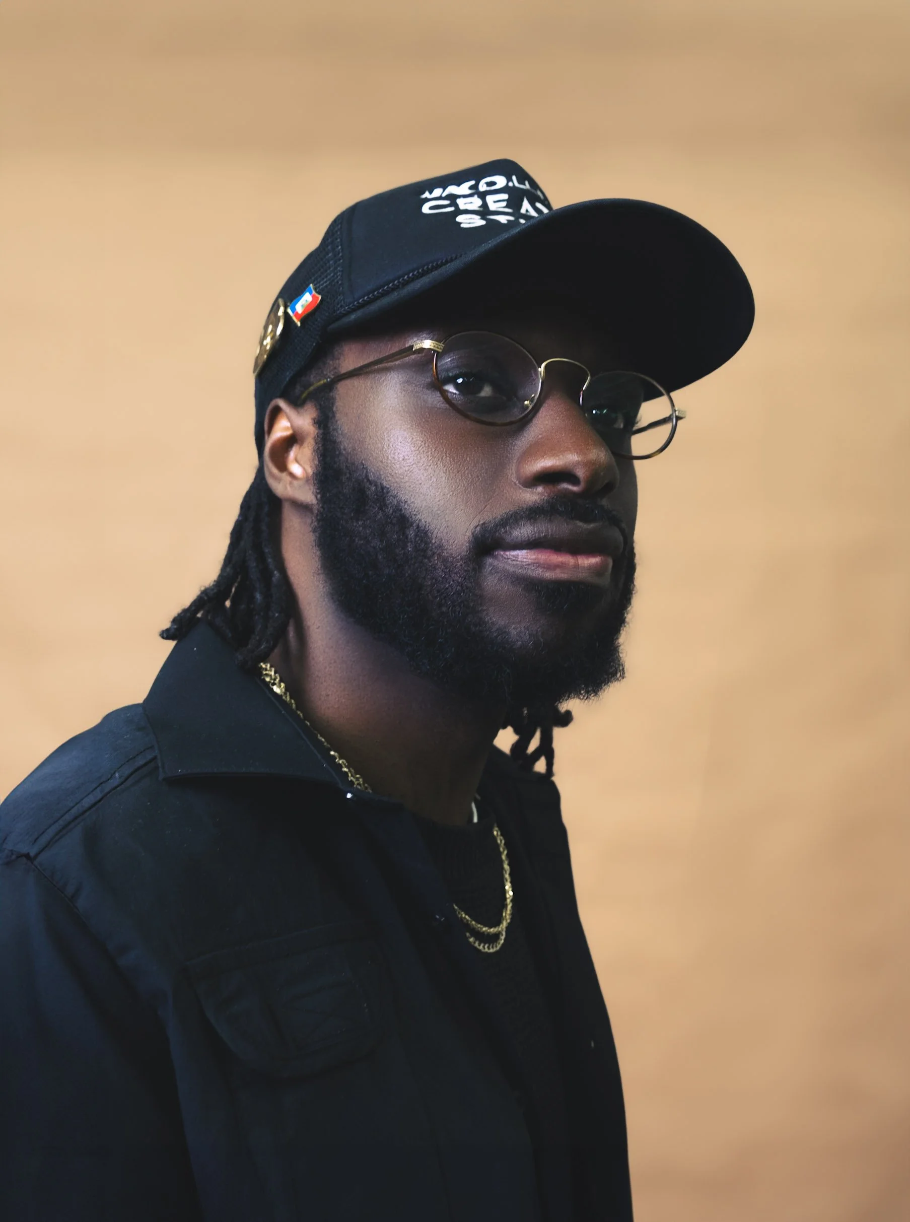 Man wearing a black cap and glasses, with a black jacket and gold chain, against a beige background.