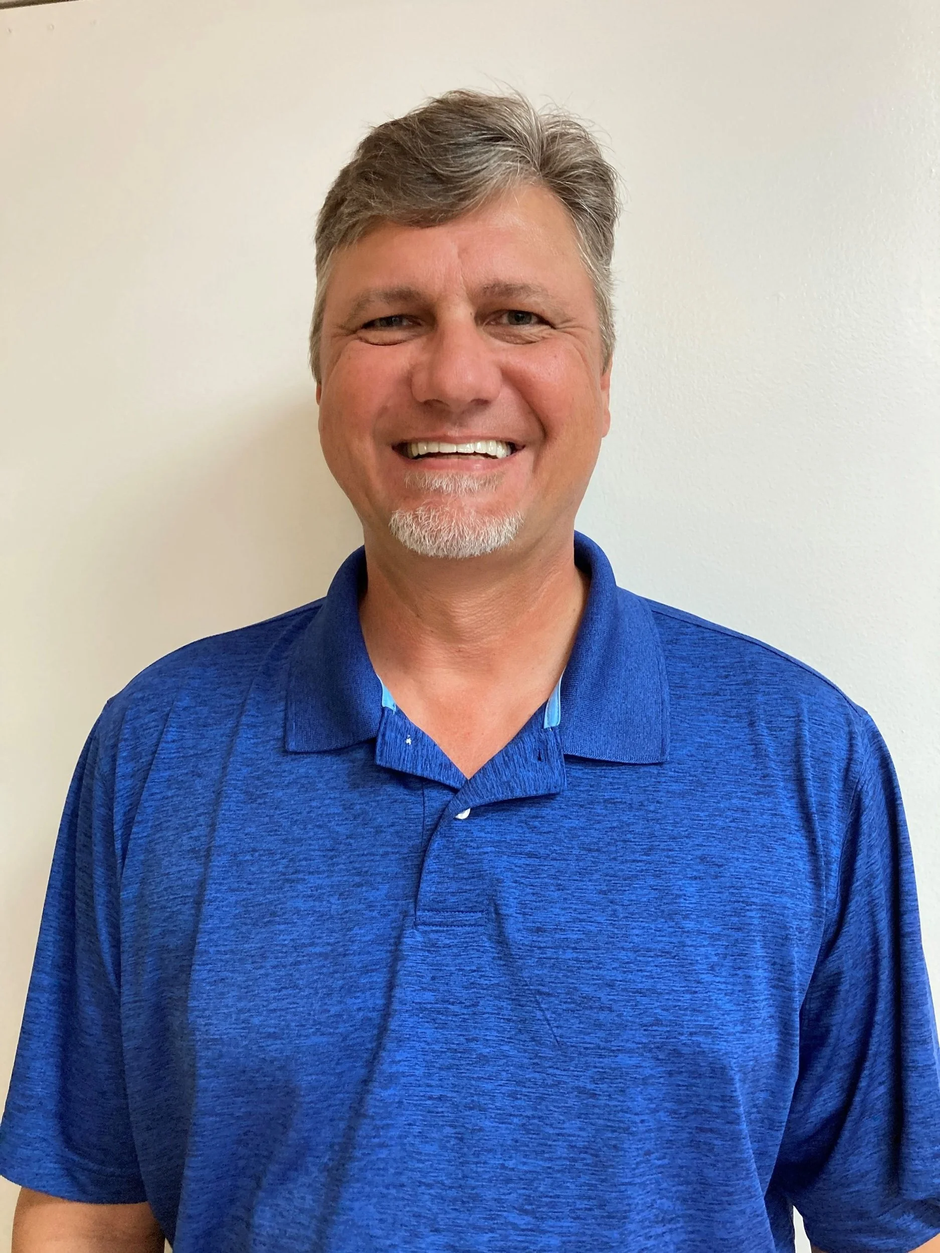 A smiling middle-aged man with tan skin, gray hair, a chin beard, wearing a blue collared shirt, standing against a plain white wall.
