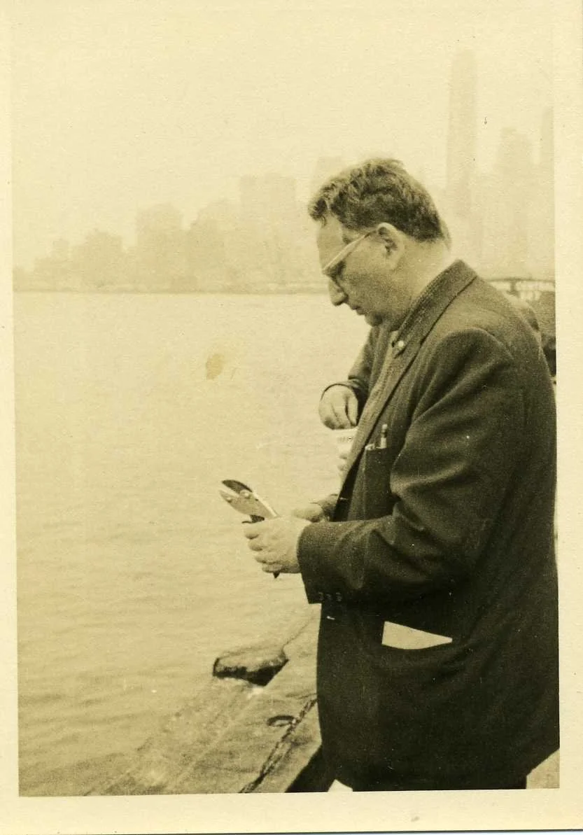 A man in a suit and glasses standing by a body of water, looking at an object in his hands, with a city skyline in the background.