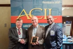 Three men in business suits at an AACIL event, with one holding a plaque, smiling for the photo.