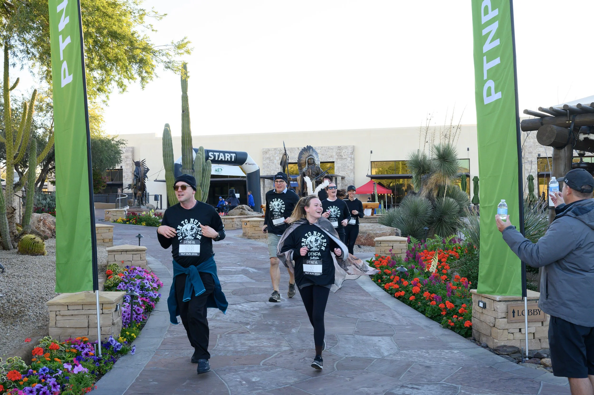 People participating in a running event outdoors, passing through a decorated arch labeled 'START', with greenery, desert plants, and flowering plants around. Some runners are smiling and wearing black shirts with event details, while a man on the ri