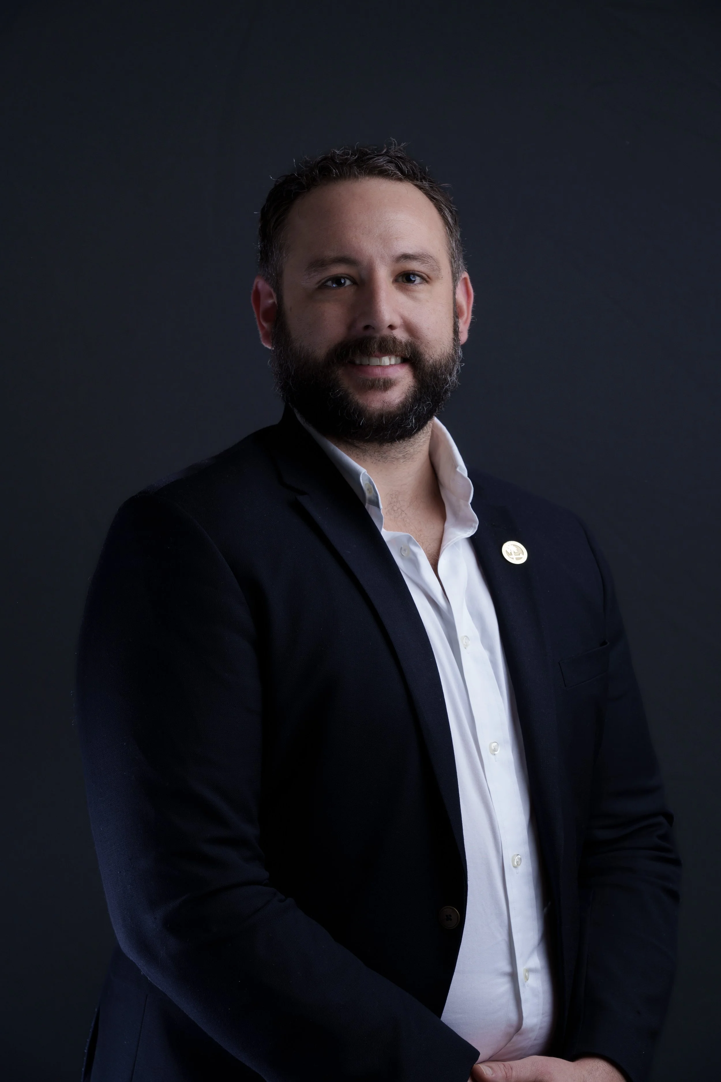 Man with dark hair and beard wearing a black blazer and white shirt, standing against a dark background.