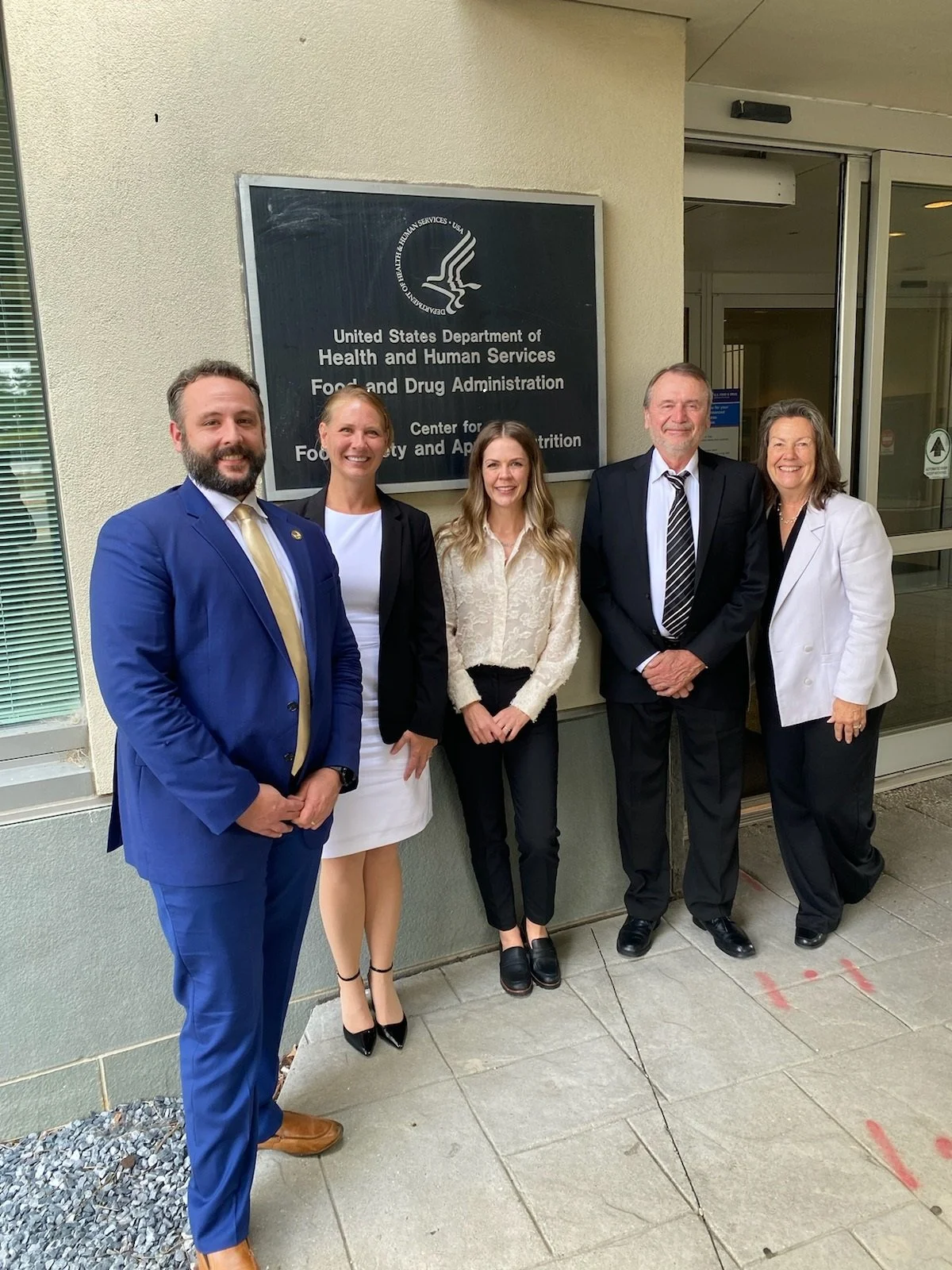 Group of five professionally dressed people standing outside the U.S. Department of Health and Human Services Food and Drug Administration building, smiling at the camera.