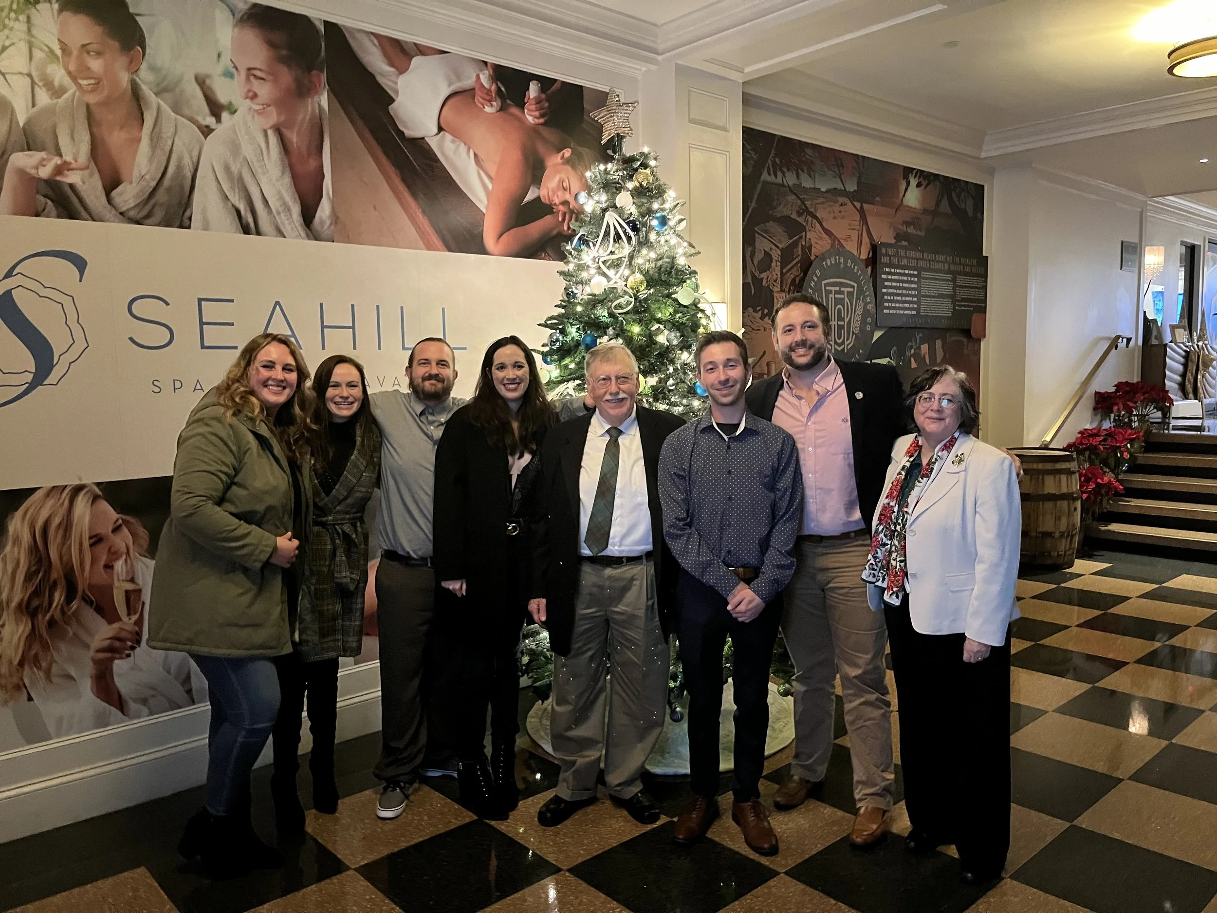 A group of eight people, including three women and five men, are standing together in front of a decorated Christmas tree at a holiday party. They are smiling and posing for the photo in an elegant indoor setting with black and tan checkered flooring