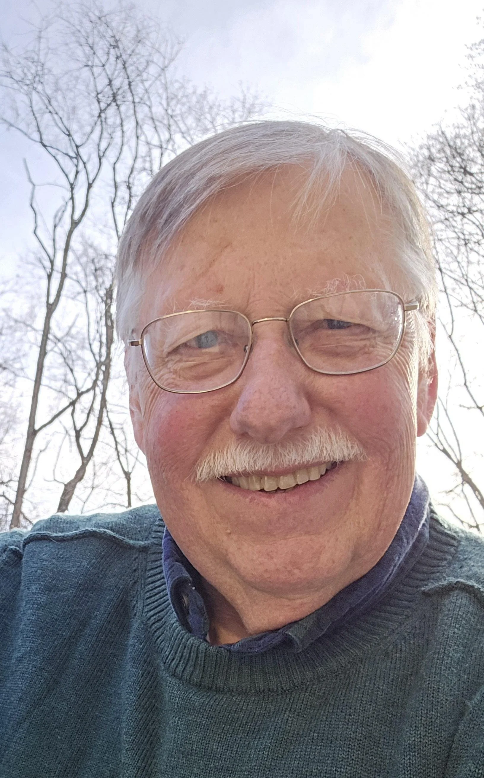 A smiling elderly man with glasses and white hair taking a selfie outdoors in front of leafless trees.