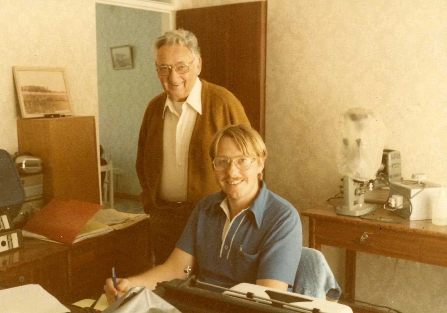 An older man with glasses and a young man with glasses sitting at a desk in an office, smiling at the camera. The office has a microscope, binders, and papers on the desk.