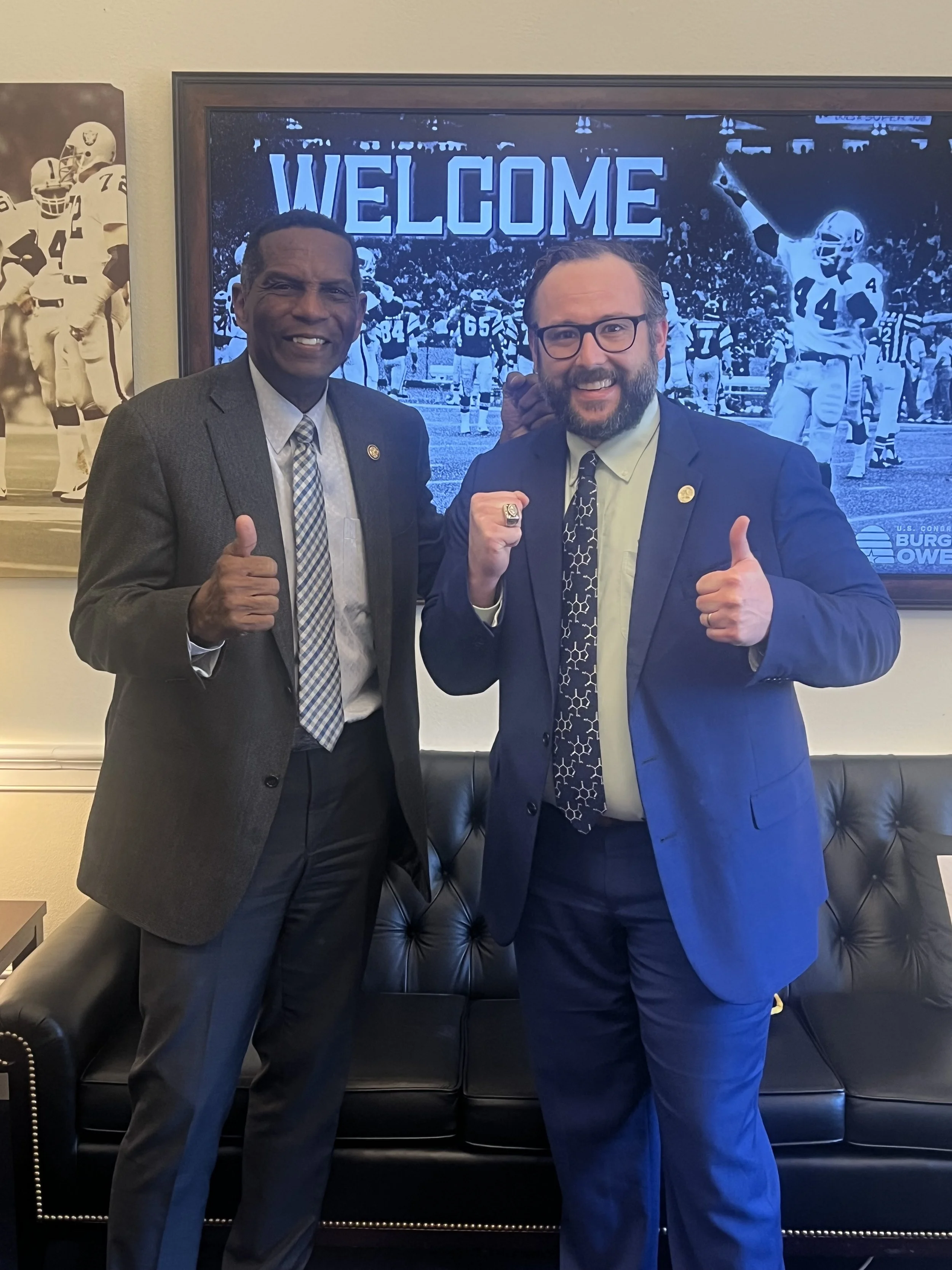 Two men in suits smiling and giving thumbs up in front of a large screen that says 'WELCOME' with a football game background.
