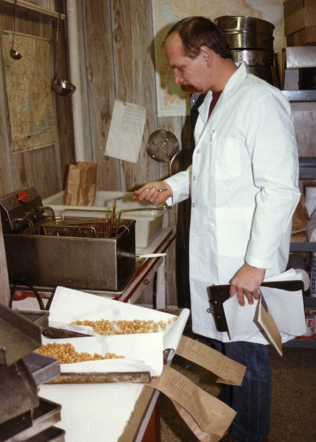 A man wearing a white lab coat working in a kitchen, frying food in a deep fryer, with trays of popcorn and paper bags on the counter.