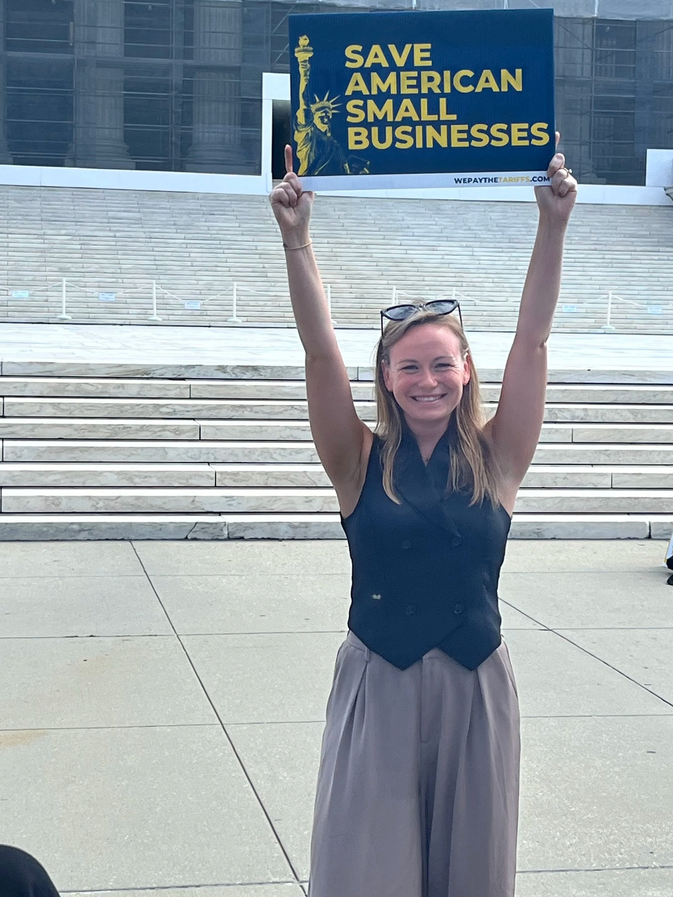 A woman smiling, holding a sign that says 'Save American Small Businesses' in front of the United States Capitol building.