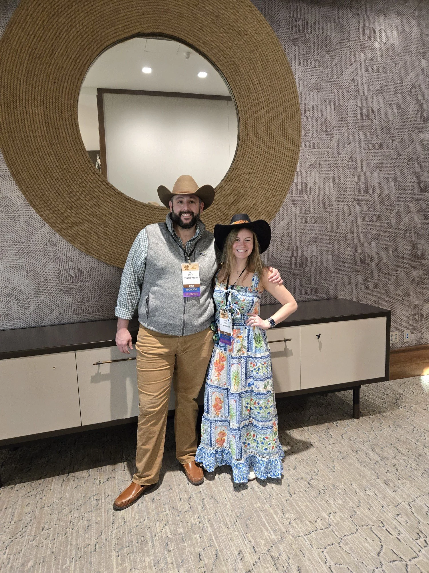 A man and woman standing in front of a large decorative mirror. The man is wearing a cowboy hat, a gray vest, khaki pants, and brown shoes. The woman is wearing a black hat, a colored patterned dress, and has a smartwatch on her left wrist. Both are 