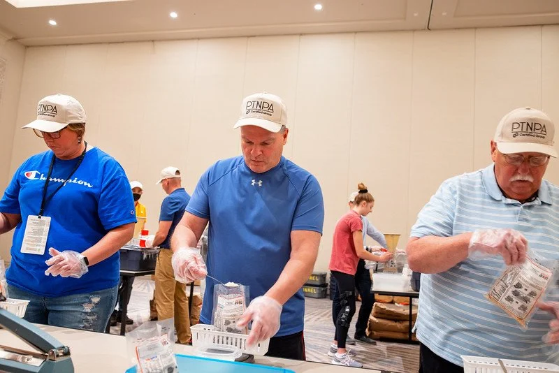 Three people wearing hats and gloves packaging food at a table in a large room, with a few other people in the background.