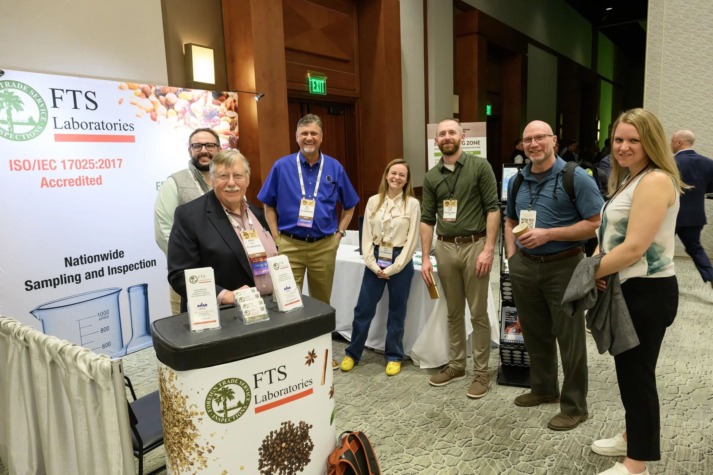 A group of seven people smiling and standing in front of a booth at a conference. The booth has signs for FTS Laboratories and features an image of a measuring beaker and grains. The background shows other conference attendees and booths.