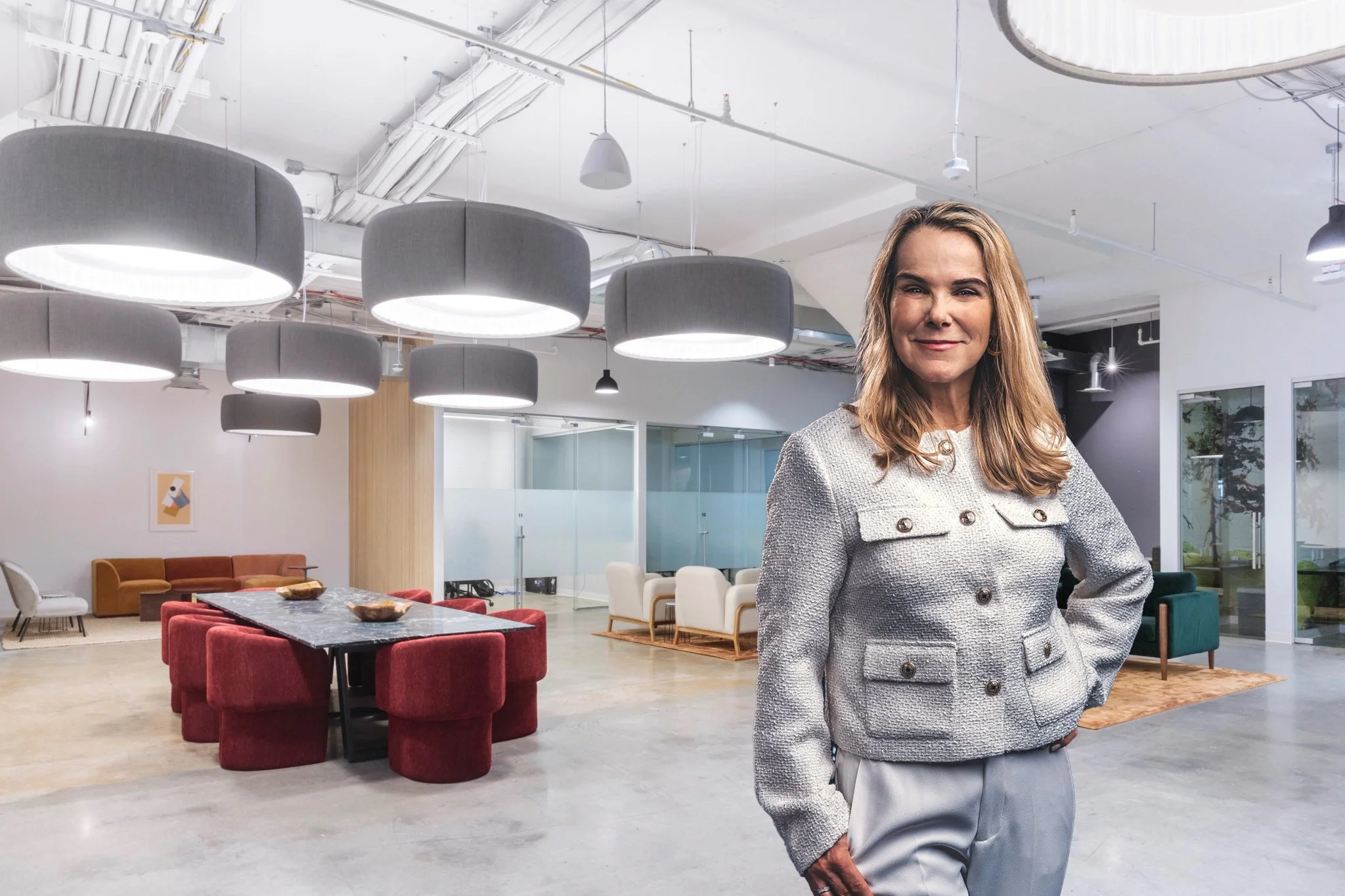 A woman with light brown hair smiling, standing in a modern, open-concept office space with contemporary furniture and large circular ceiling lights.