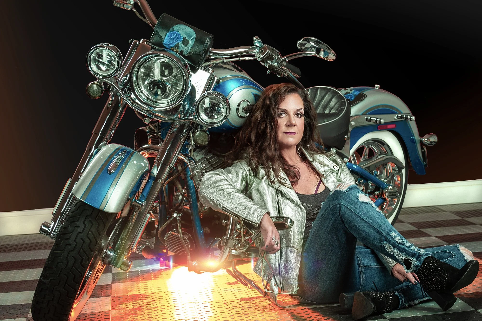 A woman with curly brown hair sitting on the ground and leaning against a large blue and chrome motorcycle with a black leather seat, wearing a silver jacket, ripped jeans, and black boots, in a studio with a dark background. Best-Fashion-Portrait-Co