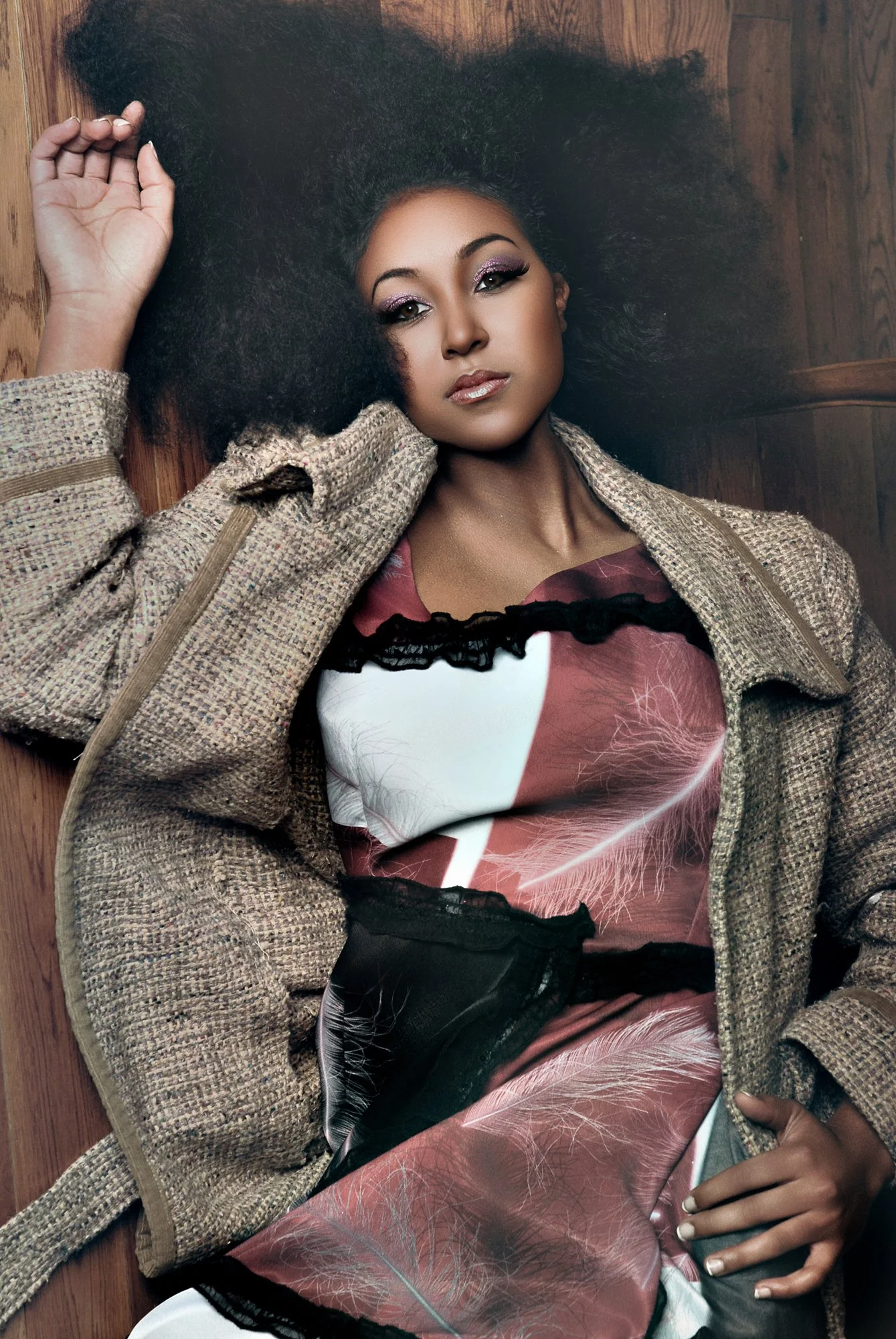 A woman lying on a wooden floor with her head resting on the ground, wearing a beige checkered jacket and a colorful dress with leaf and feather patterns, exhibiting makeup with purple eyeshadow and glossy lips. Best-Fashion-Portrait-Commercial-Busin