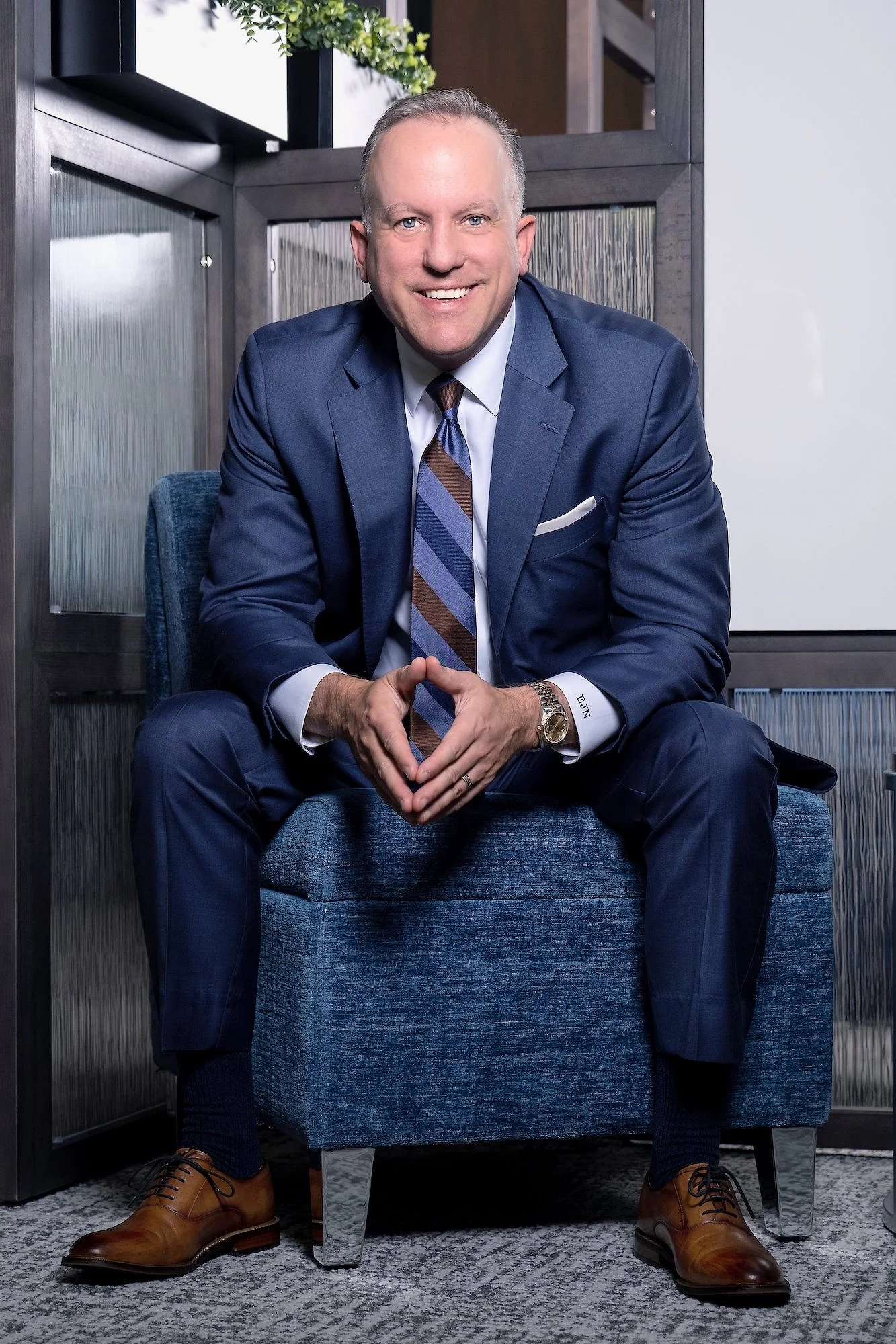 A man wearing a navy blue suit, white shirt, and striped tie, sitting on a blue chair indoors, smiling.