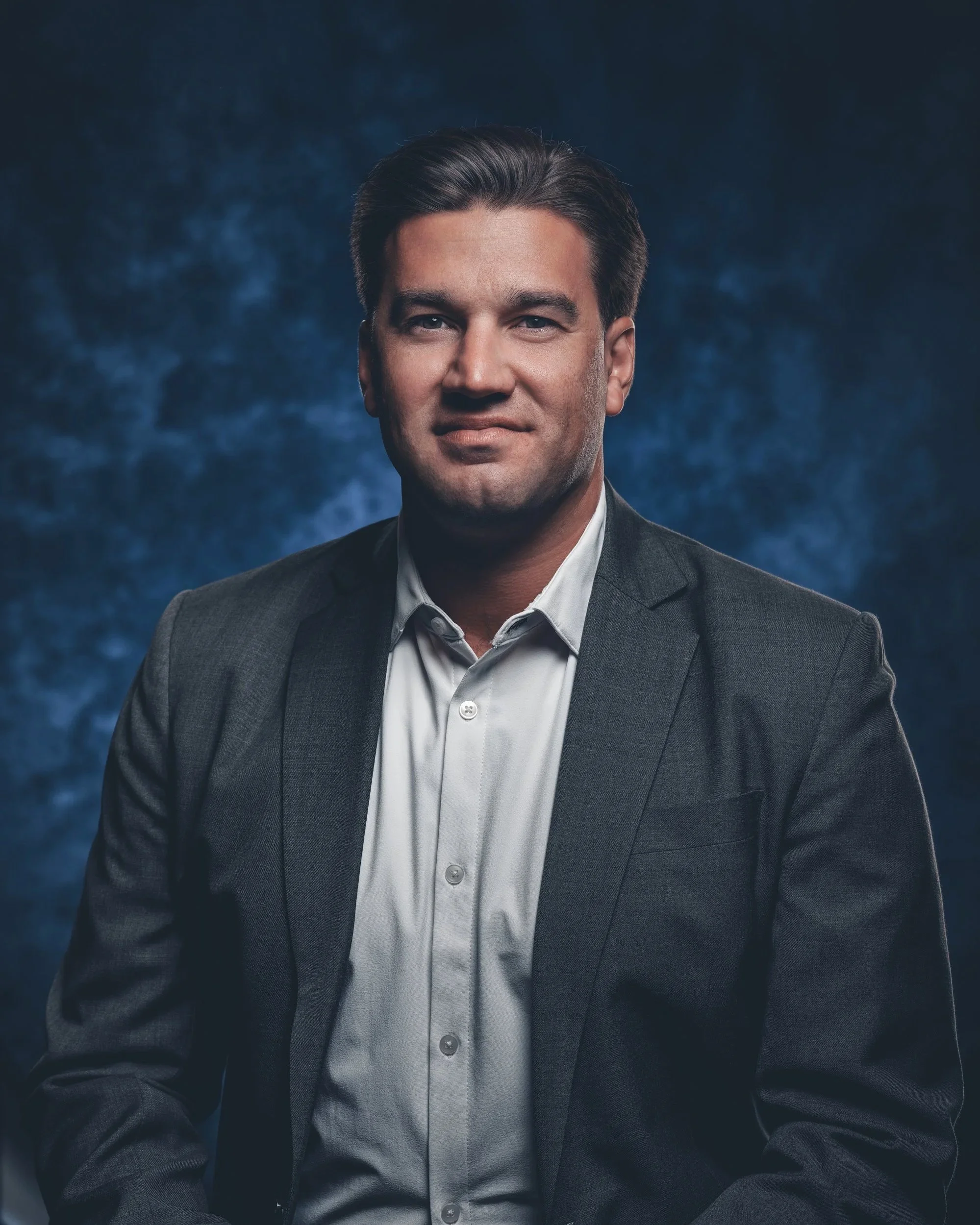 A professional man wearing a dark gray suit and white shirt, posing in front of a dark blue textured background.