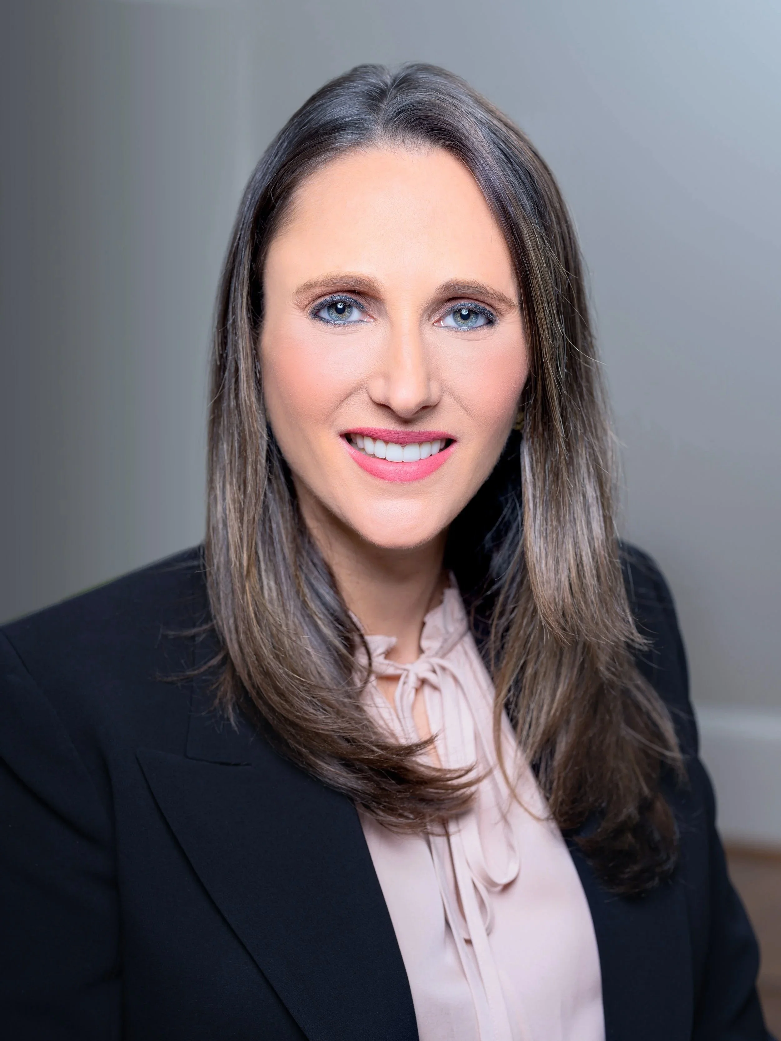 A professional woman with shoulder-length brown hair and blue eyes, smiling in a business setting, wearing a black blazer and a light pink blouse with a bow tie.