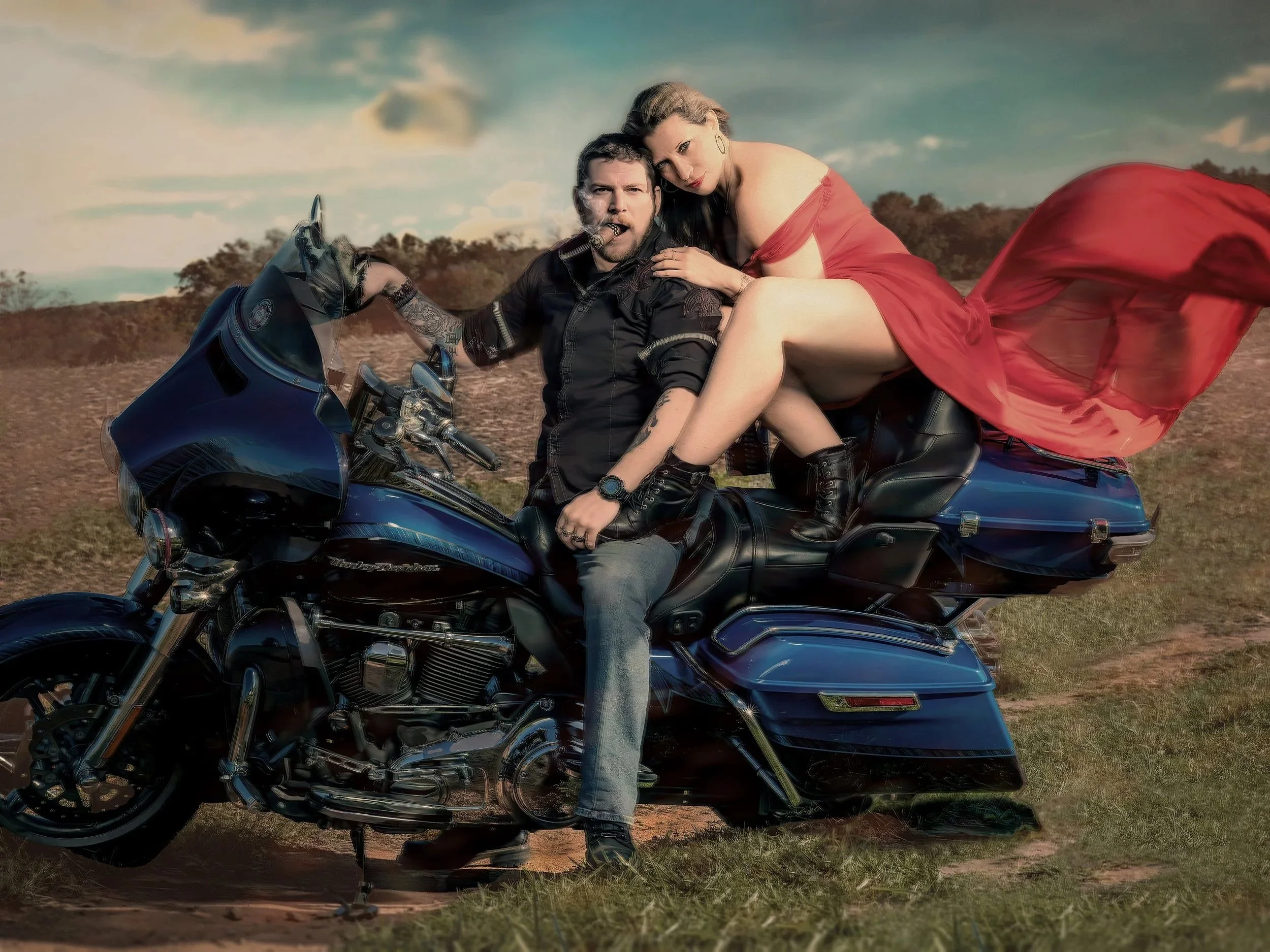 A man and woman sitting on a blue motorcycle in an open field, with the woman wearing a red dress and the man smoking a cigar.