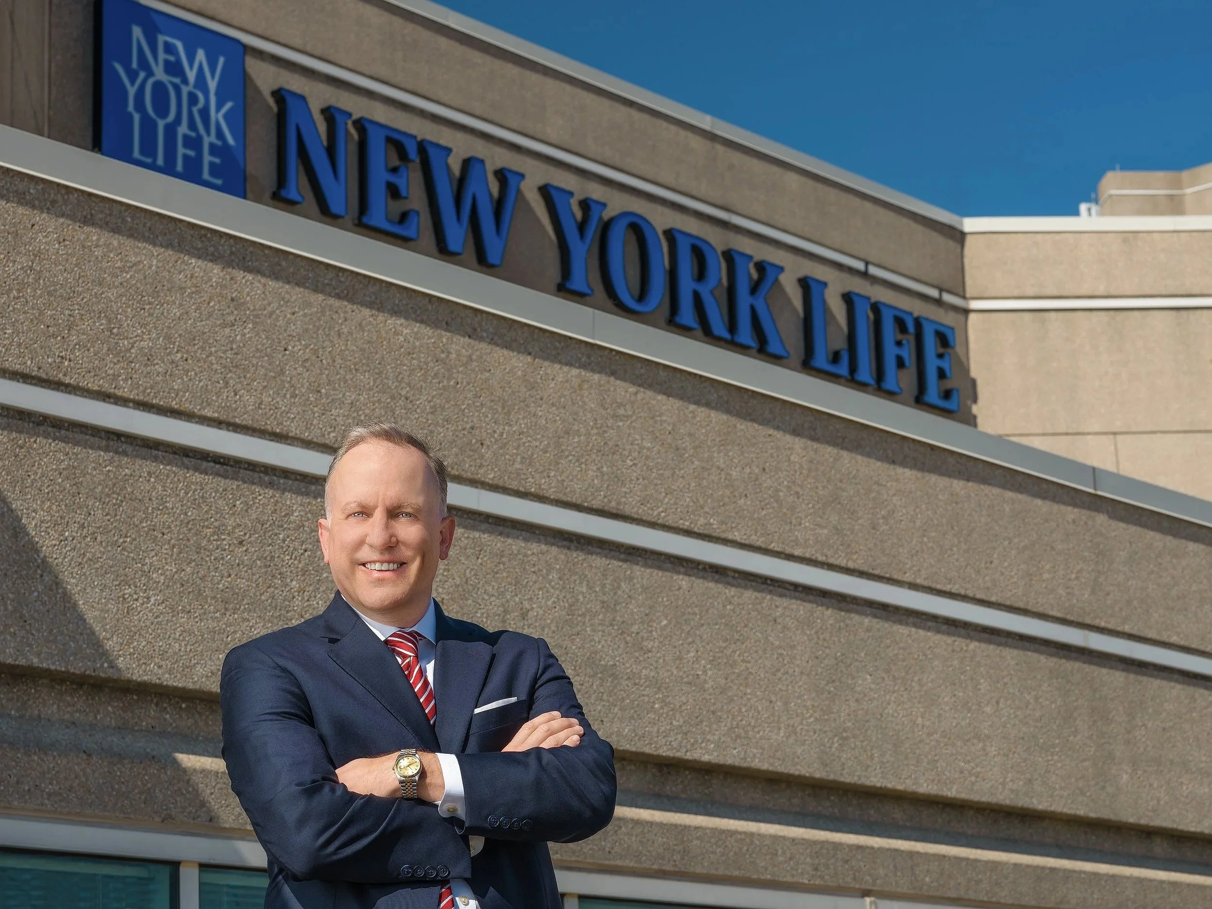 A man in a suit and red striped tie standing in front of a building with a sign that reads 'NEW YORK LIFE' and a smaller sign with the same text. He is smiling and crossing his arms.