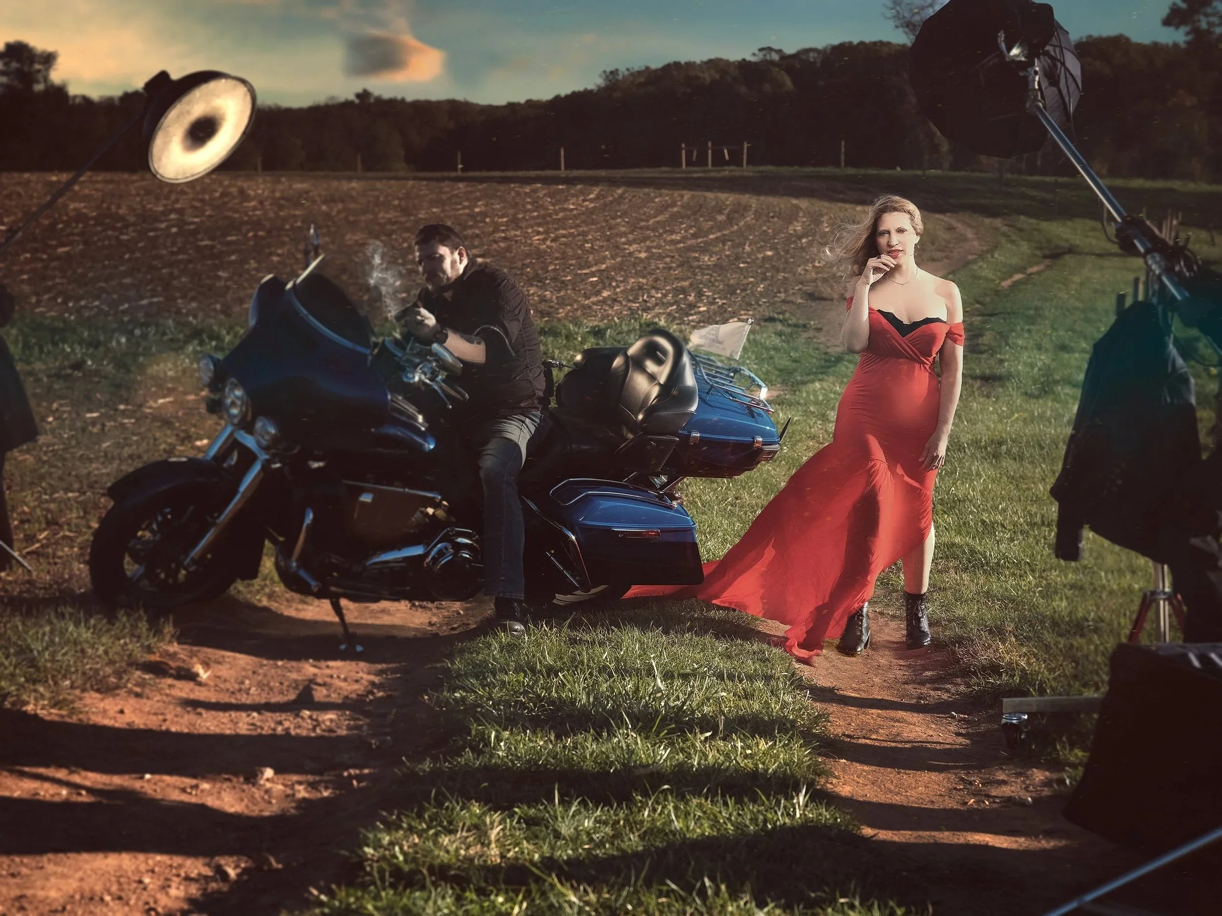 A woman in a red dress standing next to a motorcycle on a dirt path with a man sitting on the motorcycle, and professional photography equipment and lights in a rural outdoor setting. Best-Fashion-Portrait-Commercial-Business-Headshots-Corporate- pho
