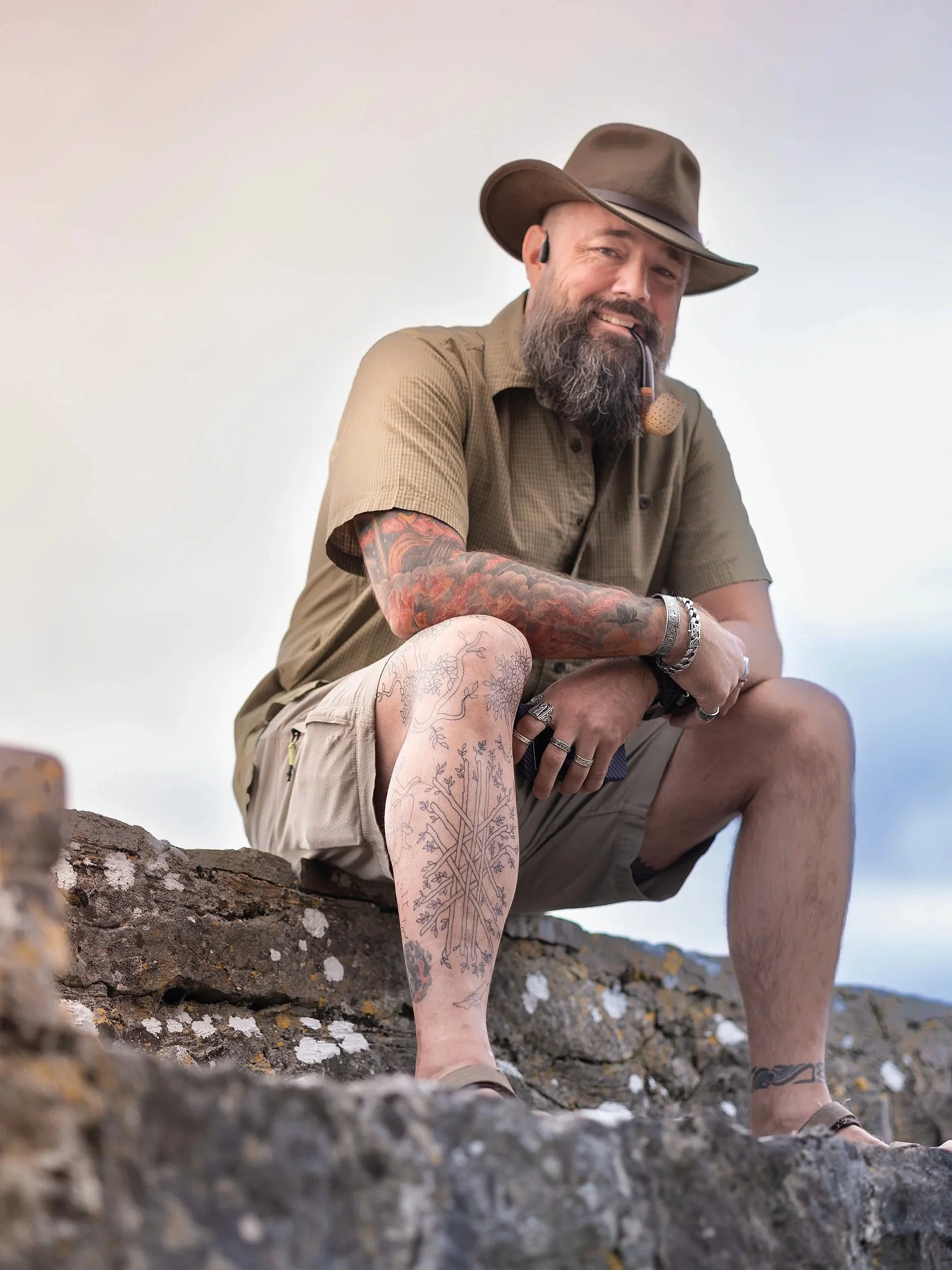 A man with a beard and tattoos, wearing a brown floppy hat, khaki shirt and shorts, sitting on rocks, smoking a pipe and smiling. Best-Fashion-Portrait-Commercial-Business-Headshots-Corporate- photography-Maryland-Washington D.C.-Pennsylvania-Virgini