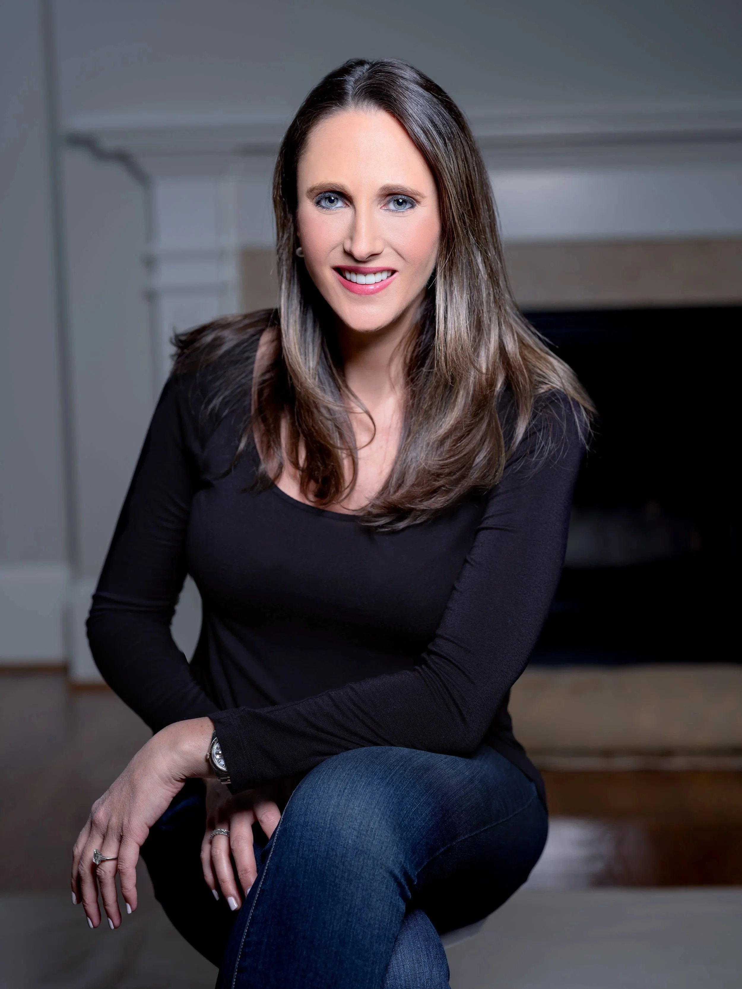 A woman with long brown hair and blue eyes, smiling, wearing a black long-sleeve top and blue jeans, sitting indoors with a fireplace in the background.  Best-Fashion-Portrait-Commercial-Business-Headshots-Corporate- photography-Maryland-Washington D