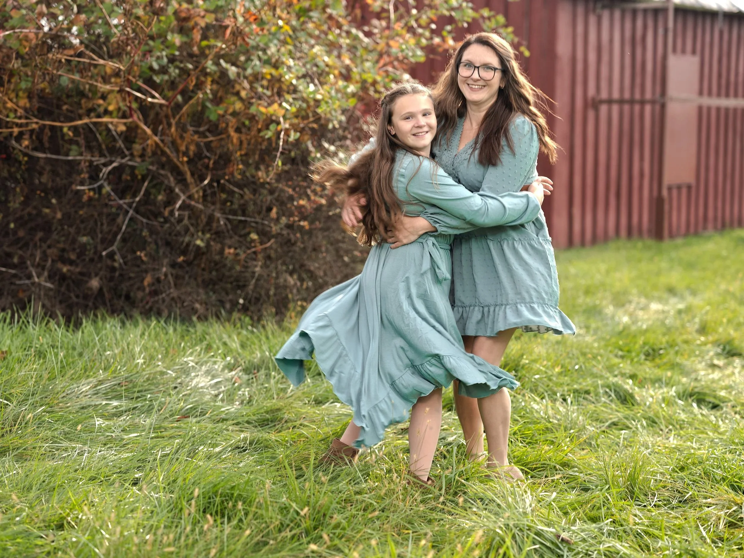Two women, one young and one older, hugging and smiling in a grassy outdoor area with a red wooden fence and bushes in the background, both wearing matching blue dresses. Best-Fashion-Portrait-Commercial-Business-Headshots-Corporate- photography-Mary